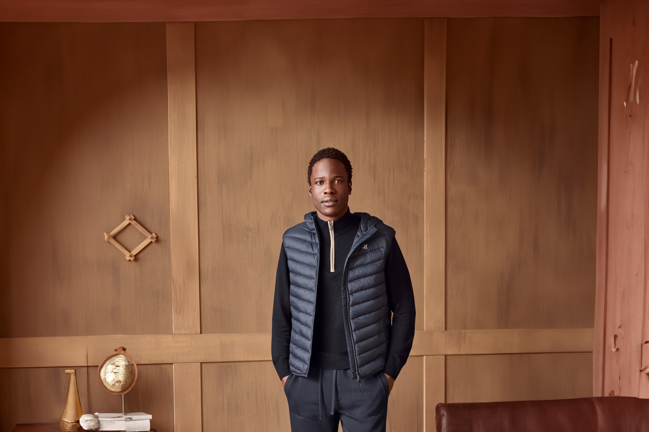 Mann in dunkelblauer Steppweste und schwarzem Rollkragenpullover, stehend vor einer Holzwand.
