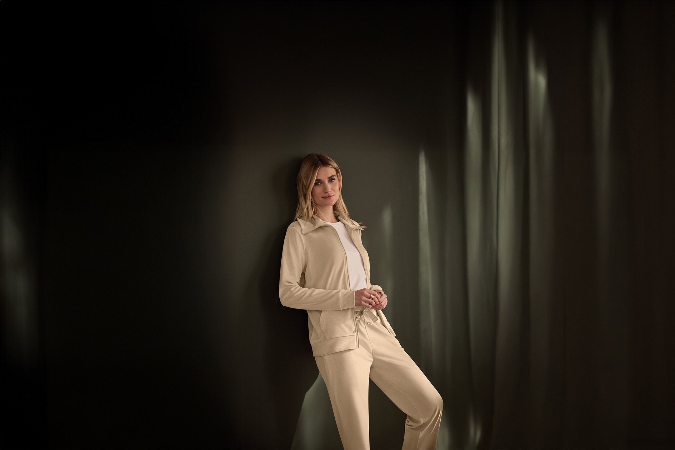 Frau in beigem Trainingsanzug und weißem T-Shirt lehnt an einer dunklen Wand.