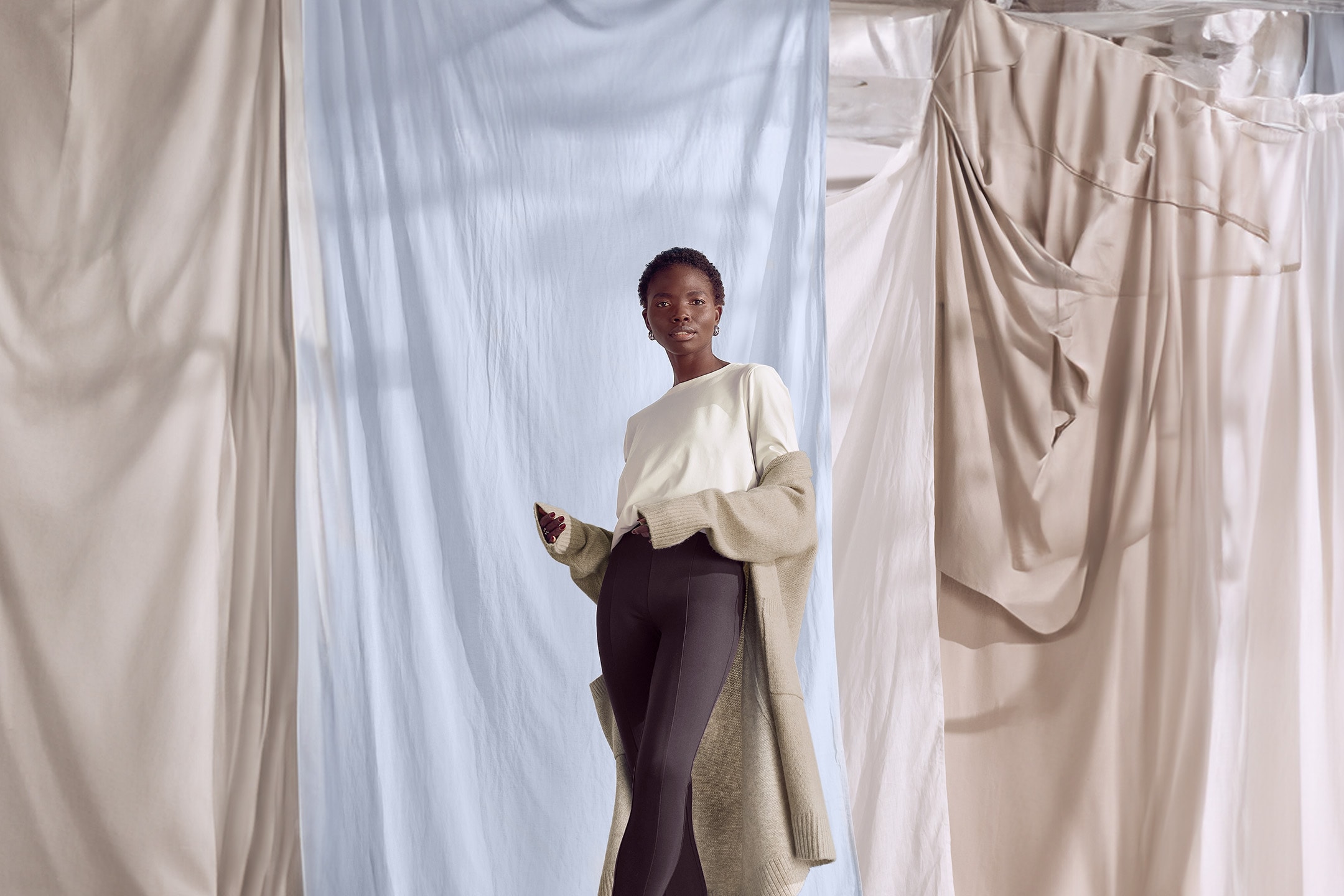 Femme en pull blanc, legging noir et cardigan beige sur fond de tissus.