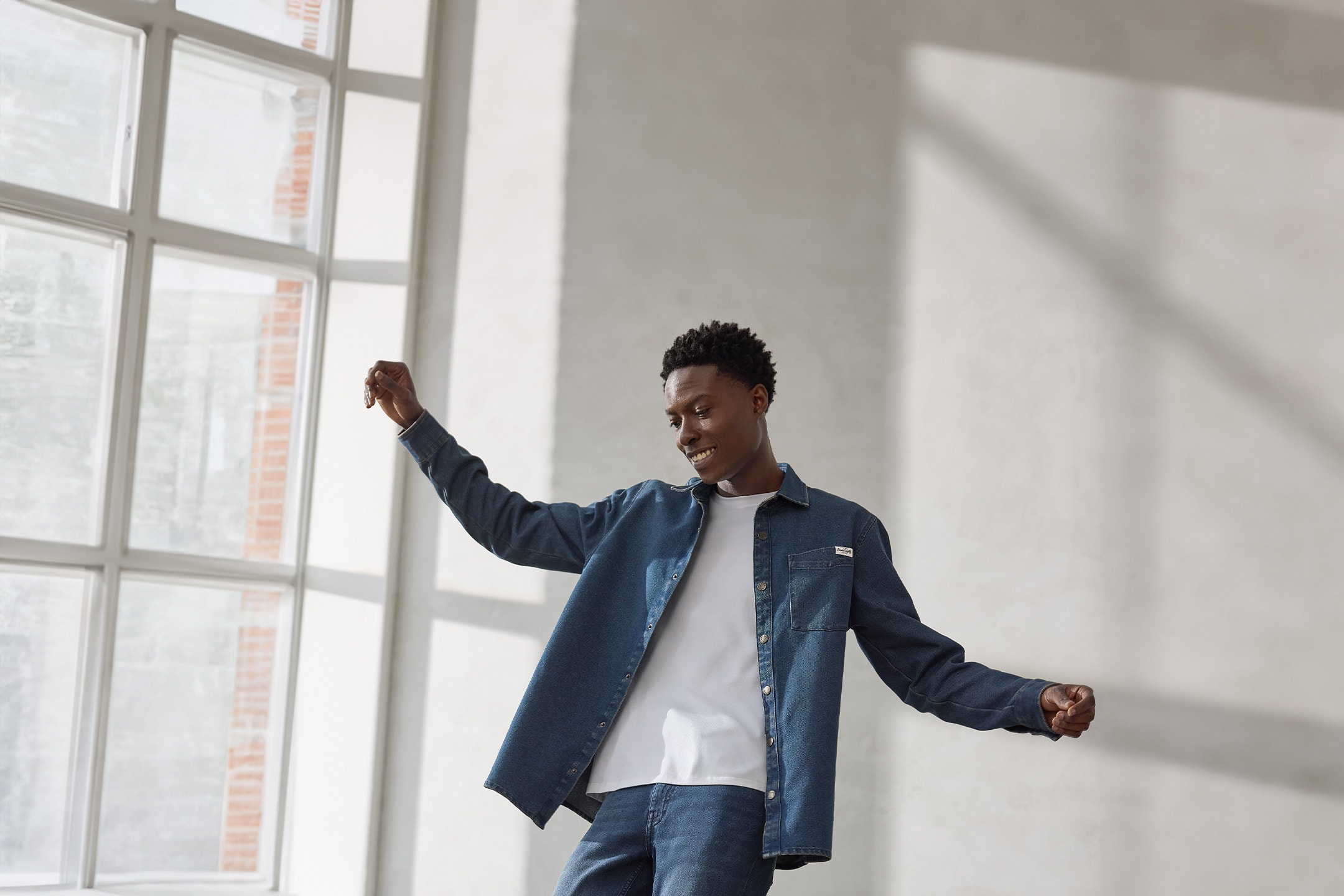 Homme en chemise en jean et t-shirt blanc dansant devant une fenêtre.