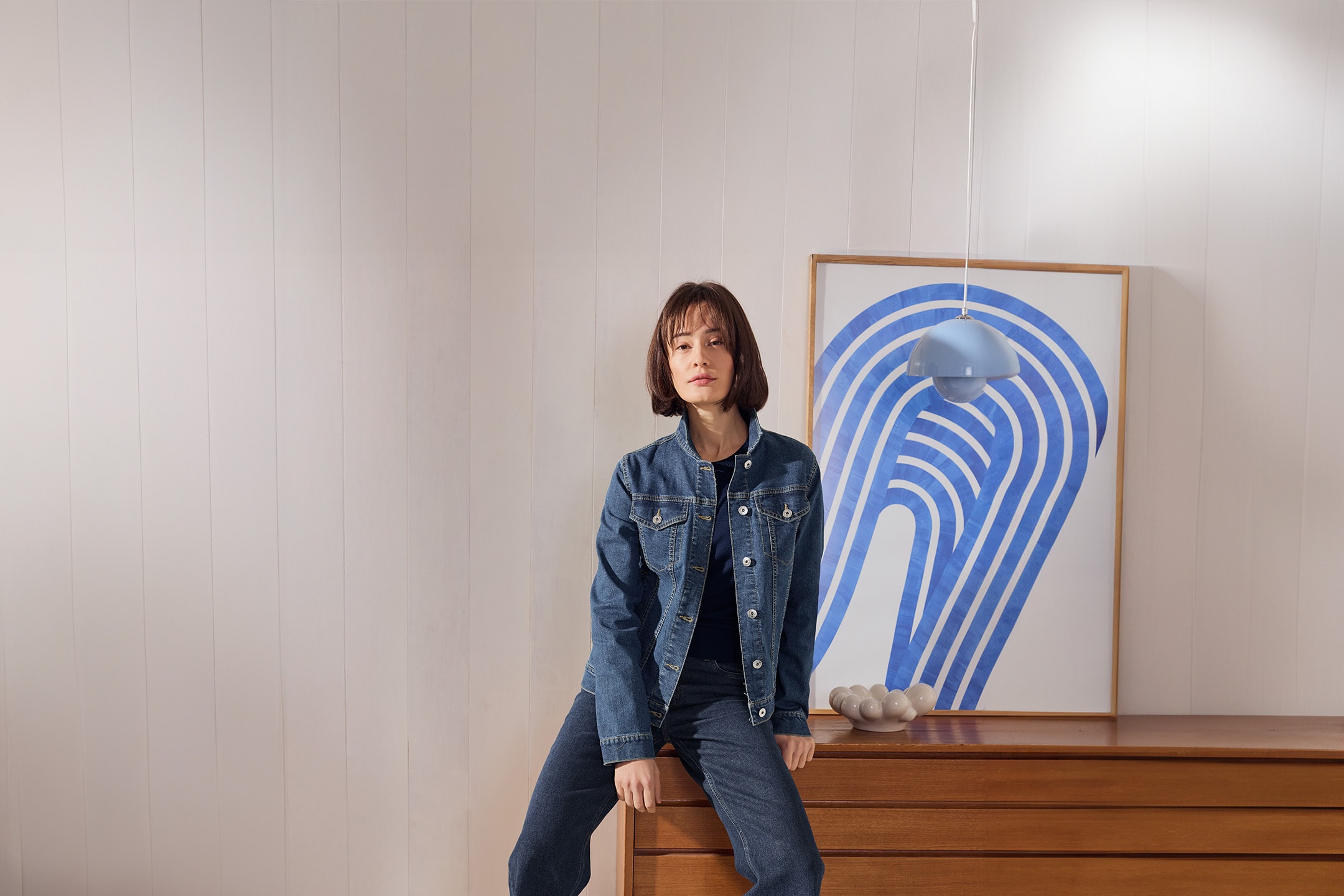 Femme en veste et pantalon en jean, assise sur une commode en bois avec un tableau abstrait.