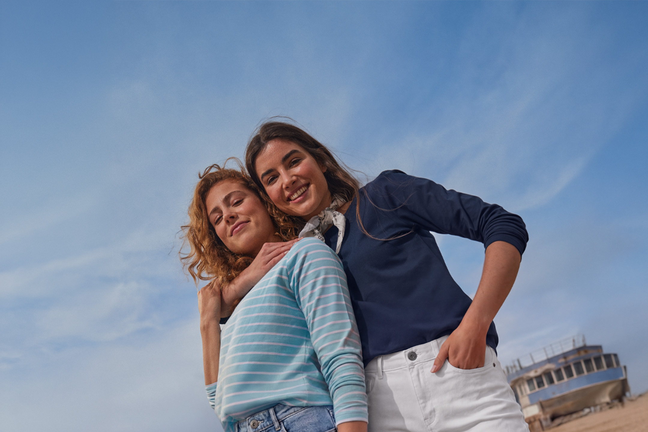 Due donne sorridenti in maglie a maniche lunghe e jeans, con una nave sullo sfondo.