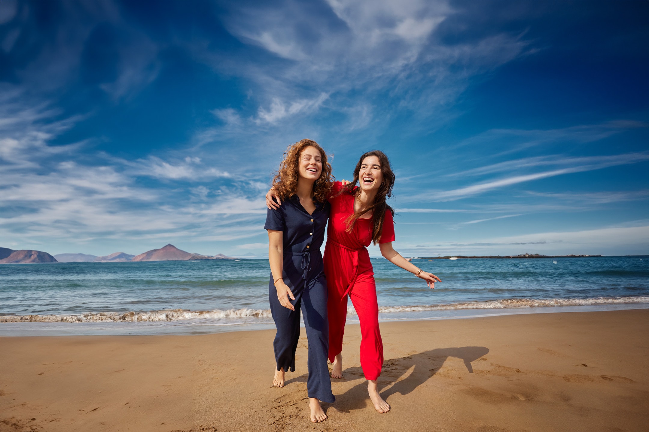 Zwei Frauen in blauen und roten Sommer-Overalls gehen lachend am Strand entlang.