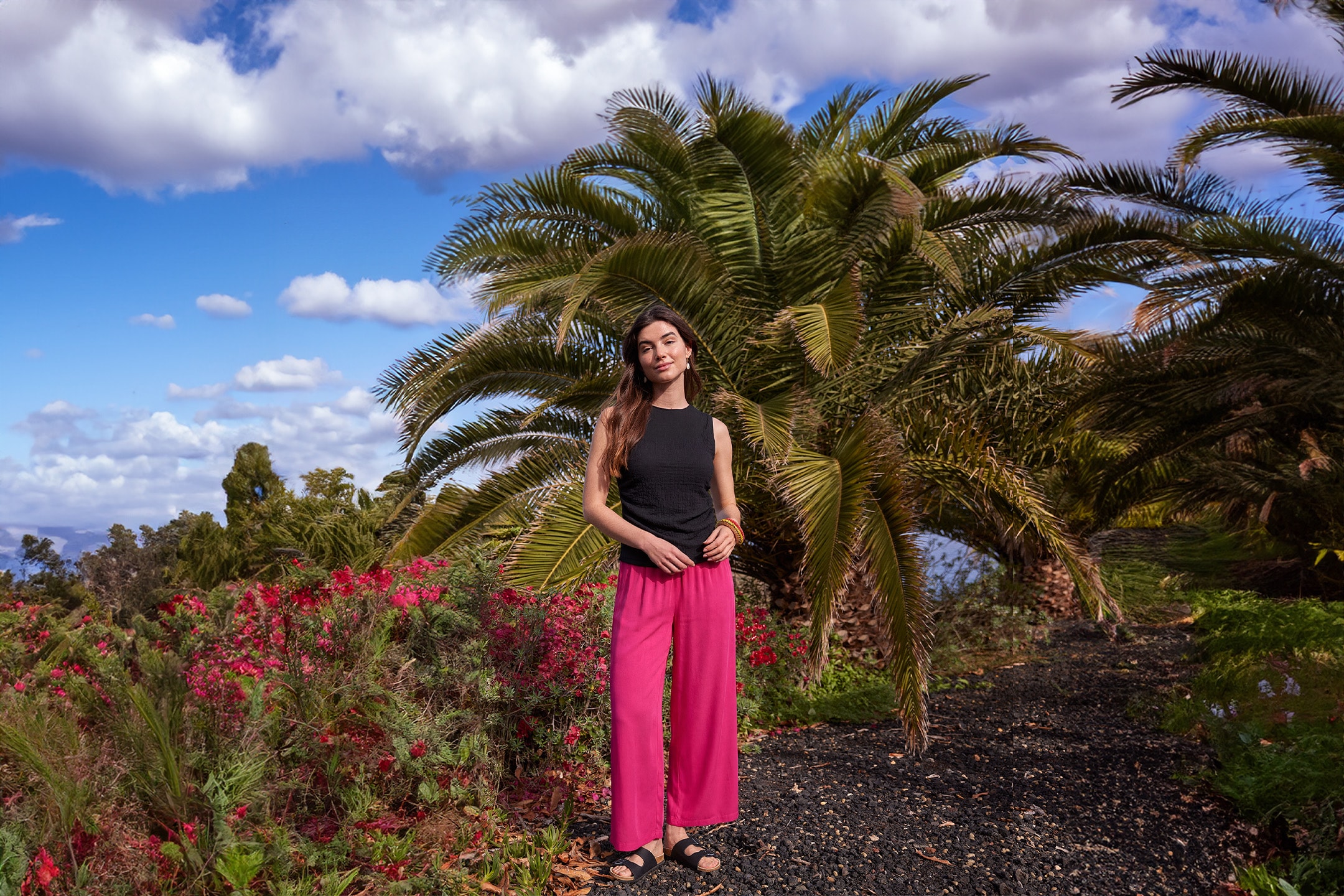 Frau in pinker Hose und schwarzem Top, steht zwischen Palmen und roten Blumen unter bewölktem Himmel.