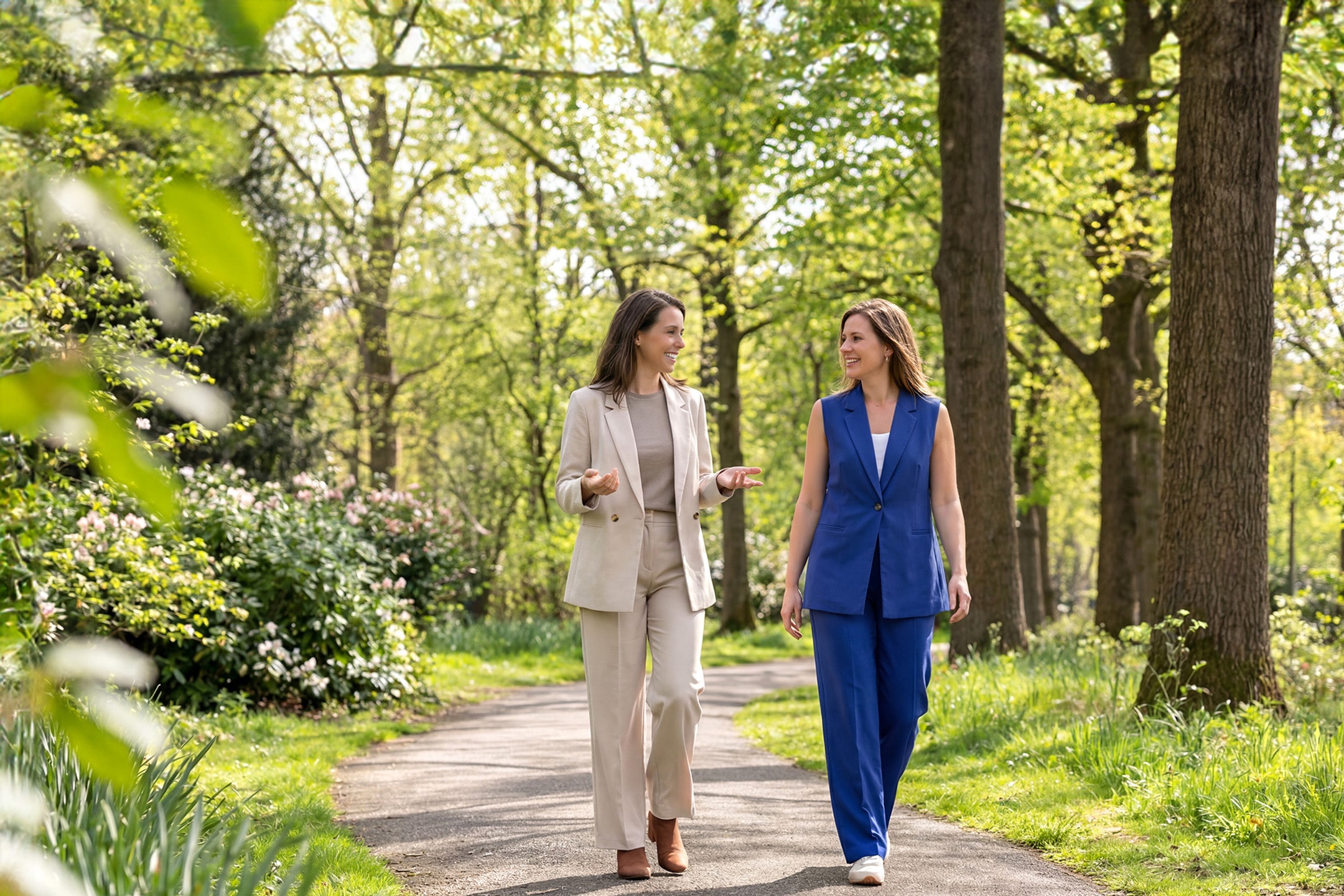 Zwei Frauen in eleganten Outfits gehen und unterhalten sich in einem sonnigen, grünen Park.