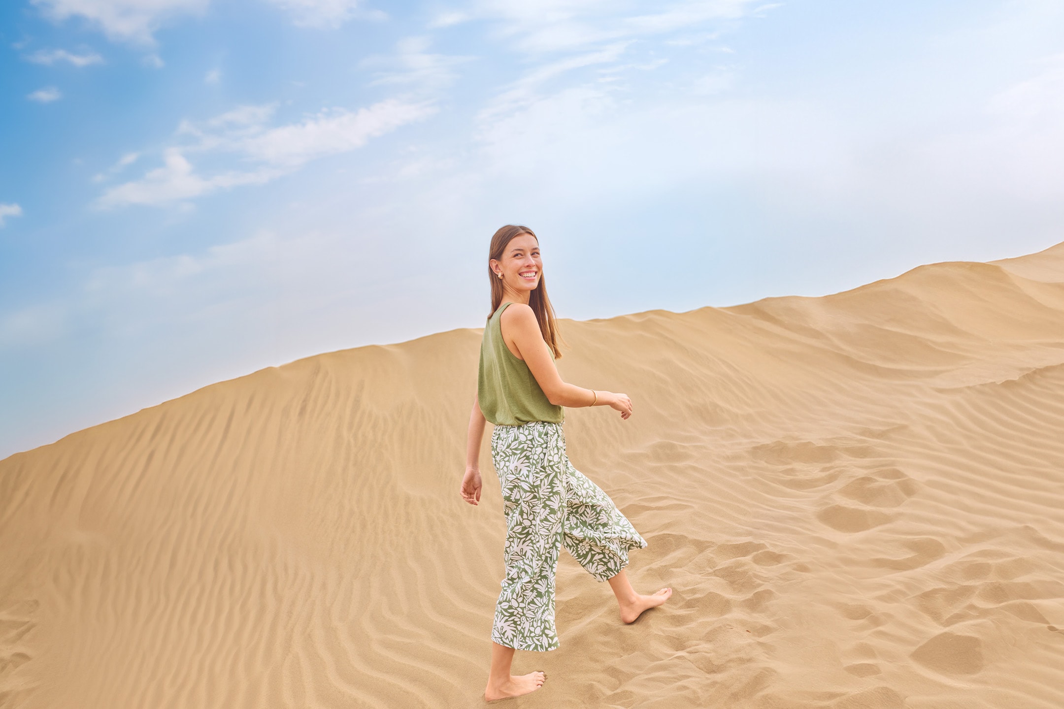 Donna sorridente in canotta verde e pantaloni a fantasia floreale, cammina a piedi nudi sulle dune di sabbia.