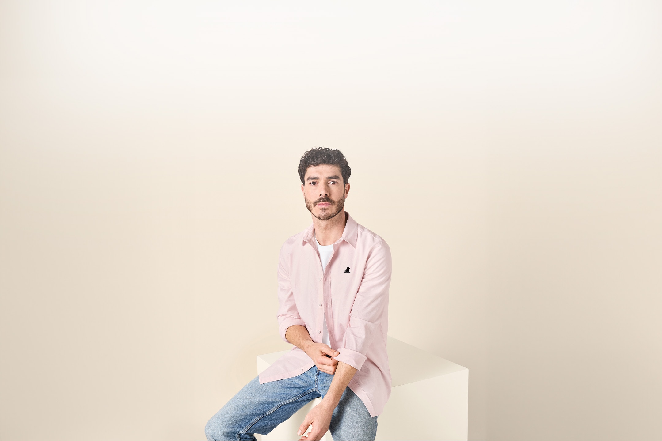 Mann in rosa Hemd und Jeans sitzt auf einem weißen Würfel