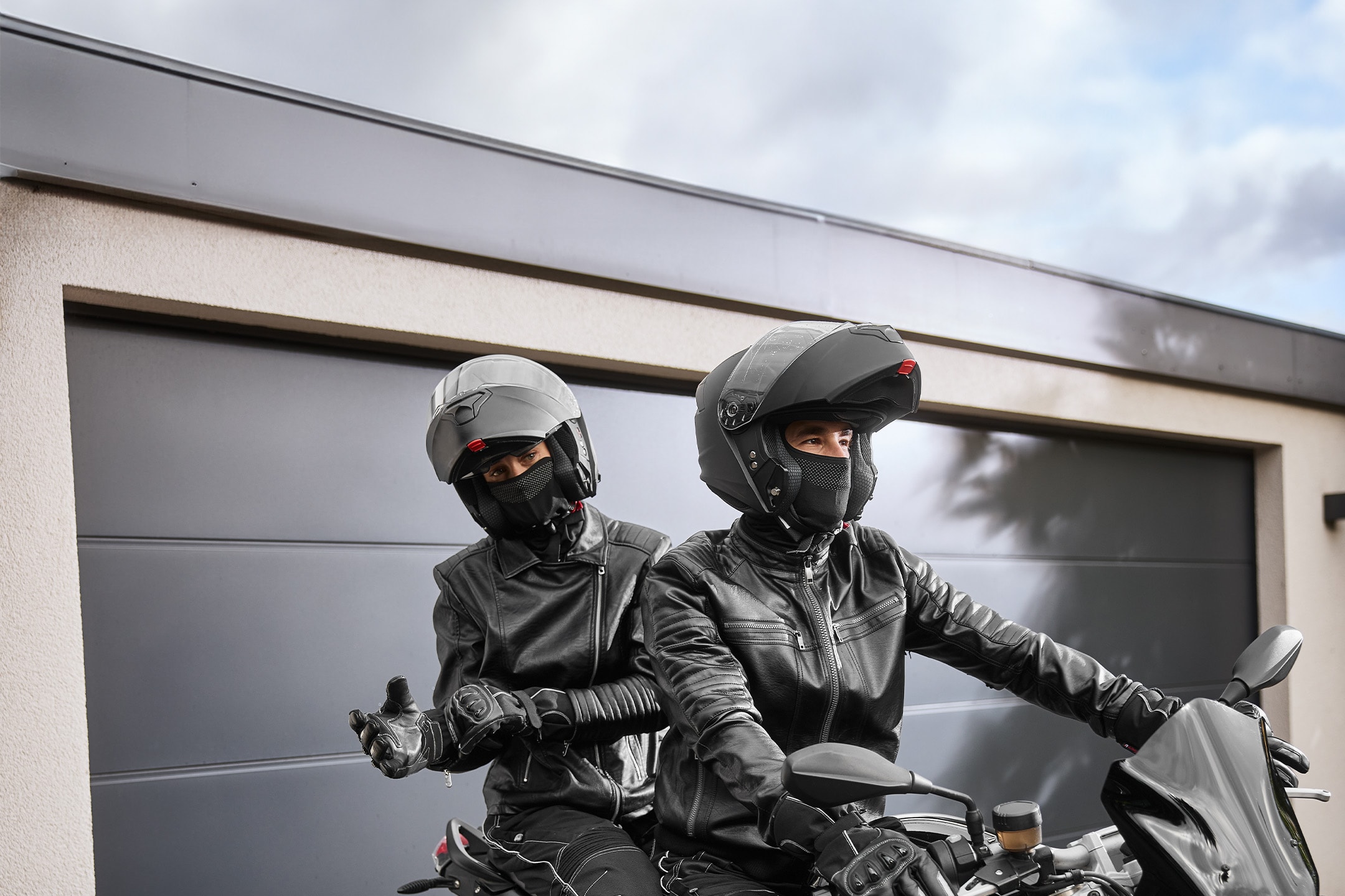 Zwei Motorradfahrer in schwarzen Jacken und Helmen, bereit zur Abfahrt auf ihrem Motorrad.