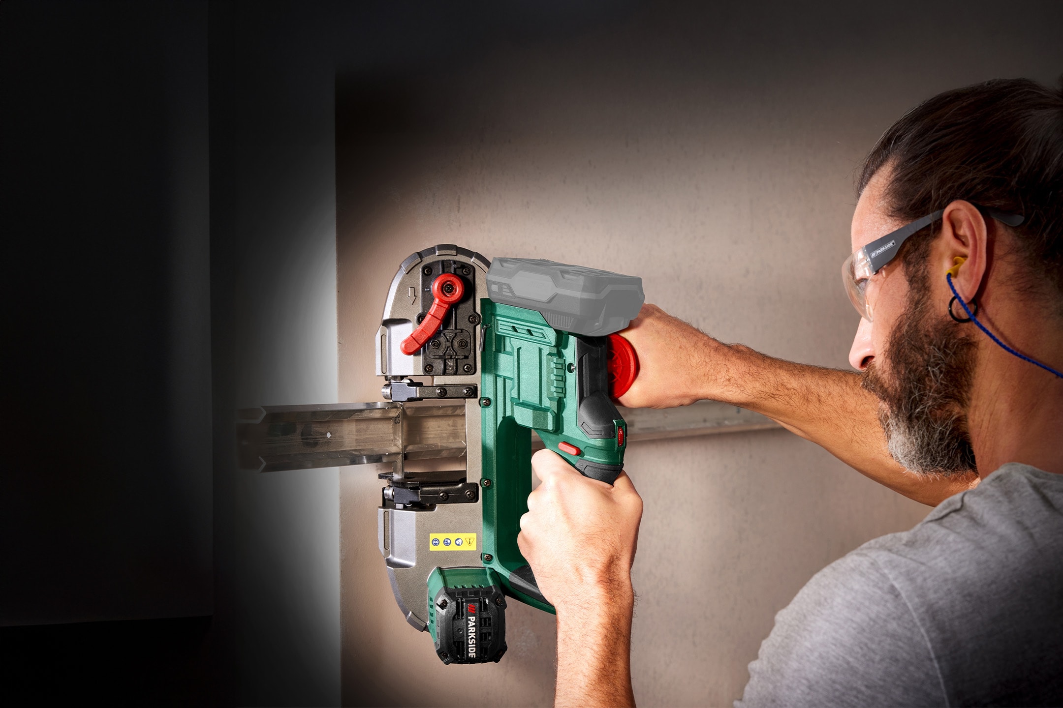 Mann schneidet Metallprofil mit einer Akku-Bandsäge, trägt Schutzbrille und Gehörschutz.