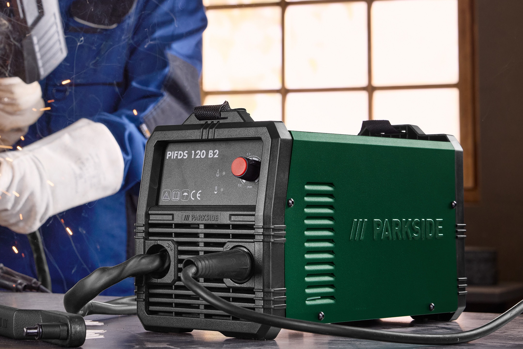 Poste à souder compact vert et noir sur une table, avec un soudeur en combinaison bleue en arrière-plan.