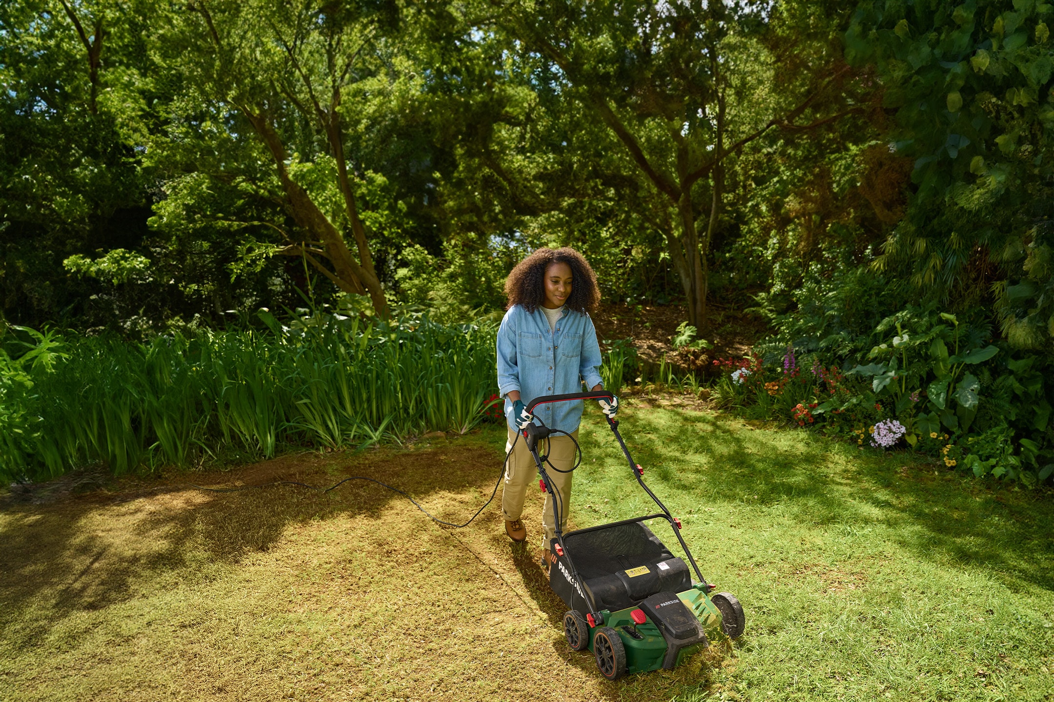 Eine Frau benutzt einen elektrischen Vertikutierer auf einem Rasen, umgeben von Bäumen und Grün.