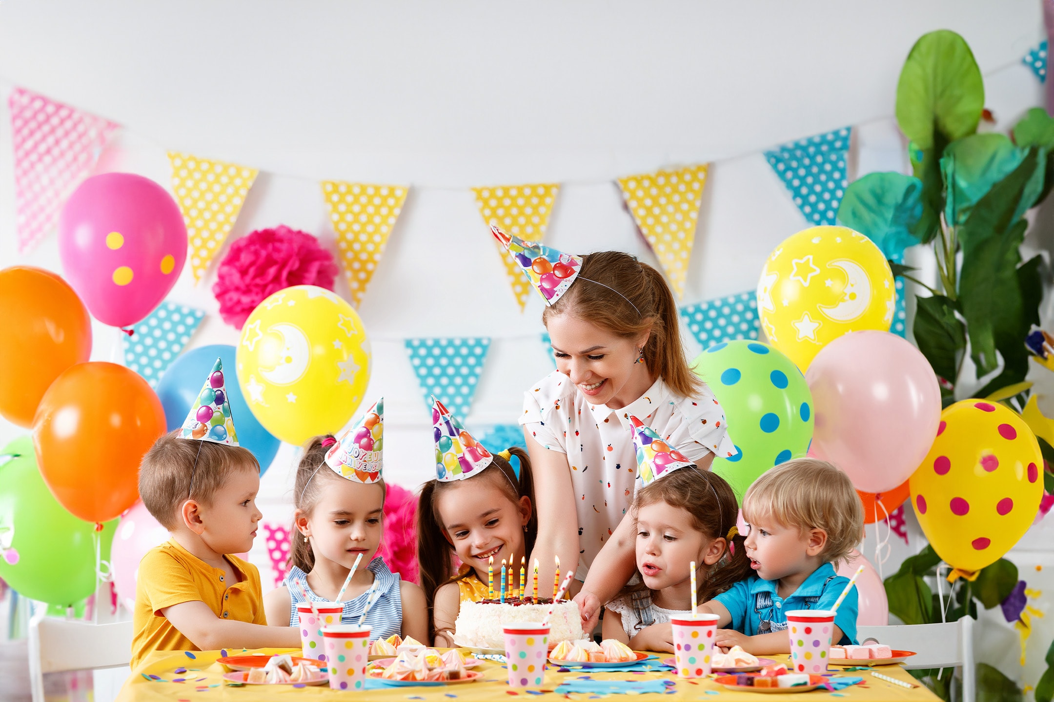 Lächelnde Kinder und Erwachsene um einen Geburtstagskuchen mit Kerzen und Ballons.