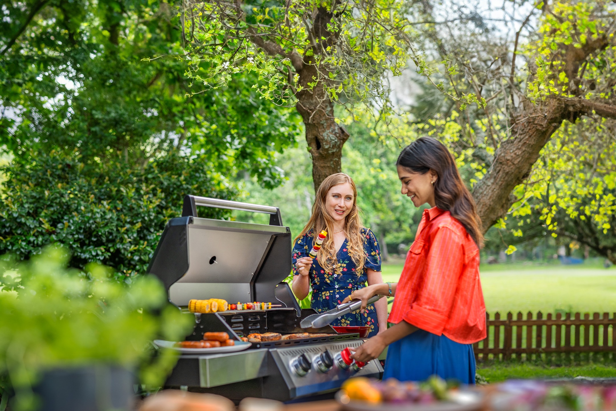 Zwei Frauen grillen Spieße, Würstchen und Mais auf einem Grill im Garten.