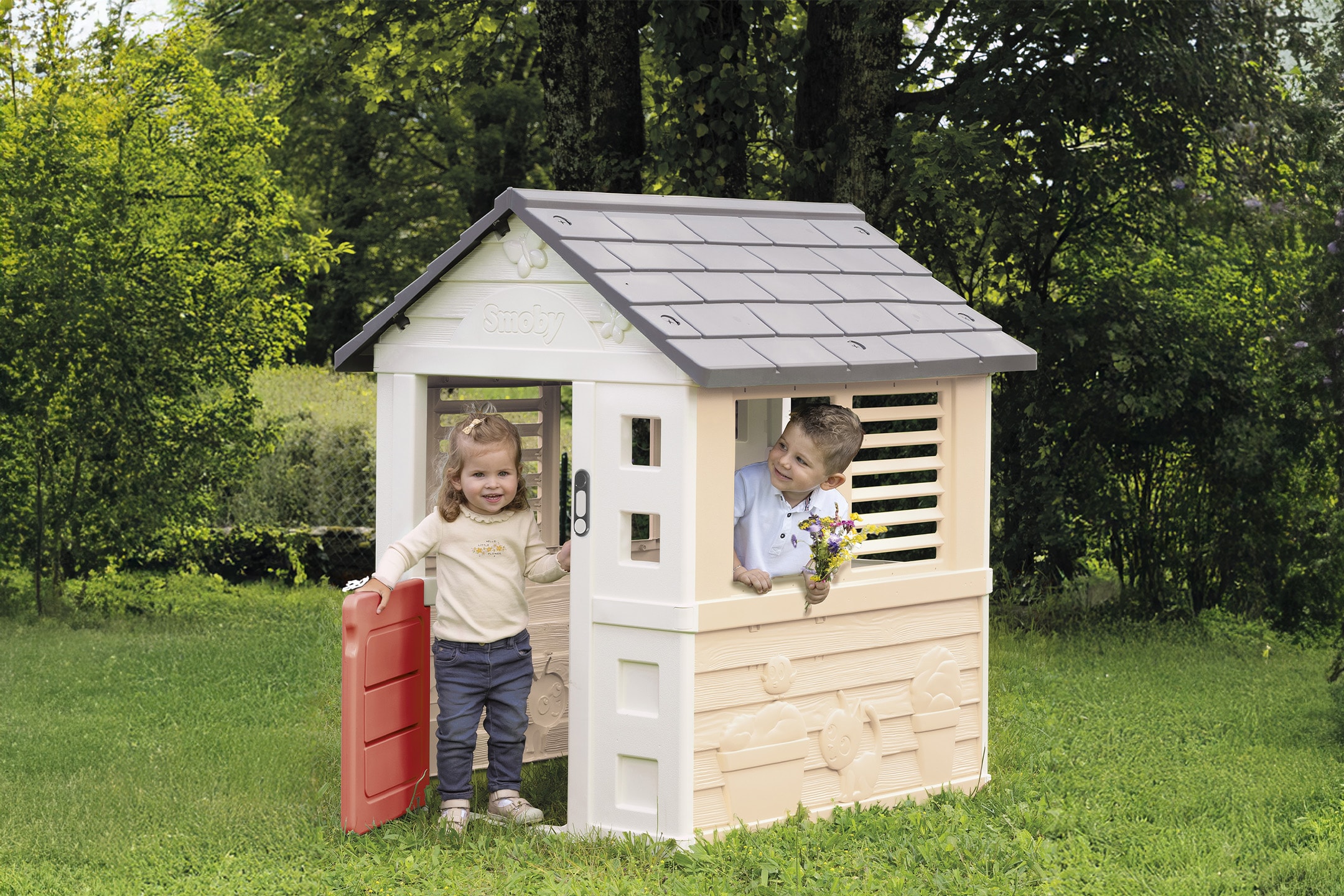 Zwei Kinder spielen in einem Gartenhäuschen aus Kunststoff, umgeben von Grün.