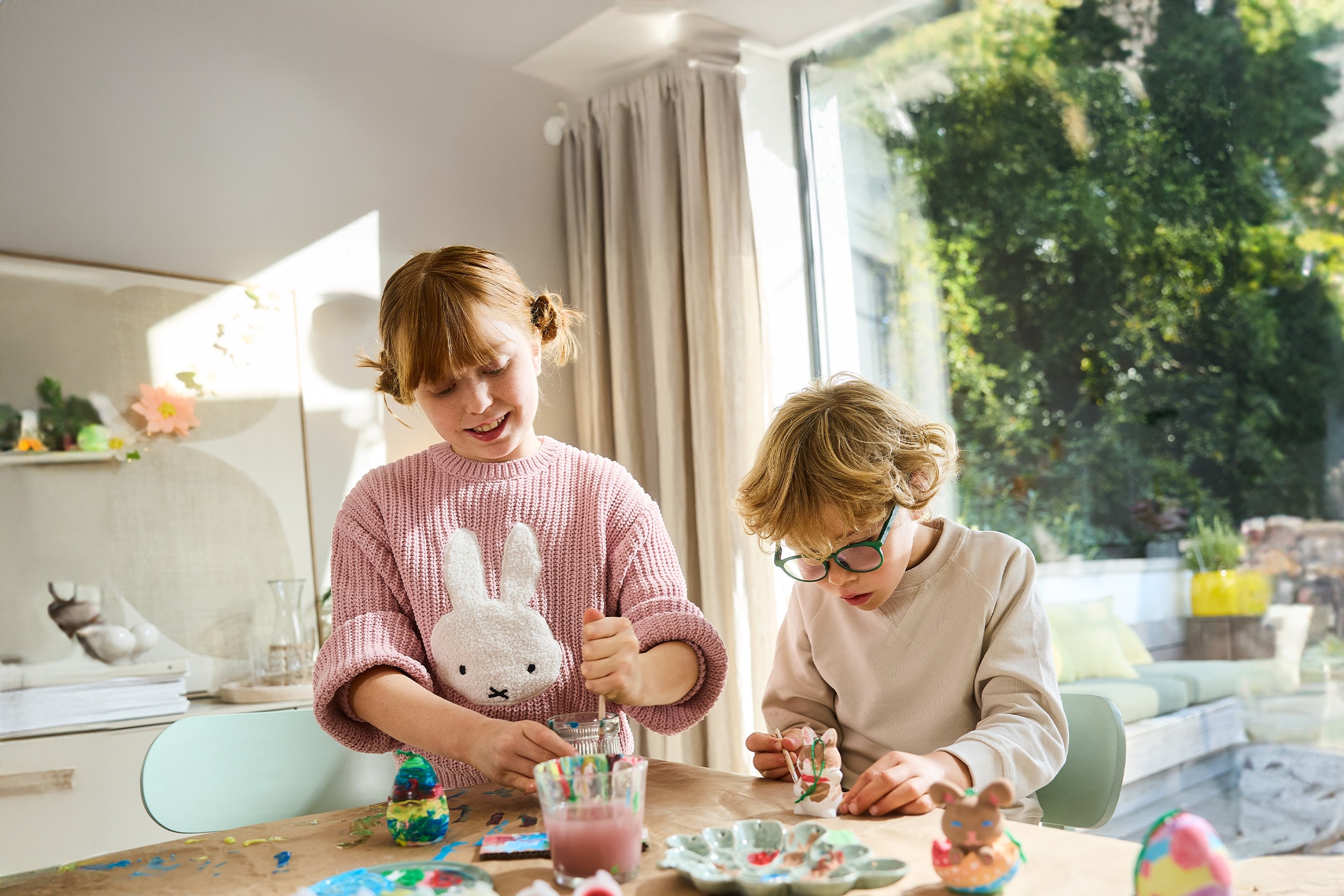 Zwei Kinder bemalen Ostereier und Hasenfiguren mit bunten Farben.