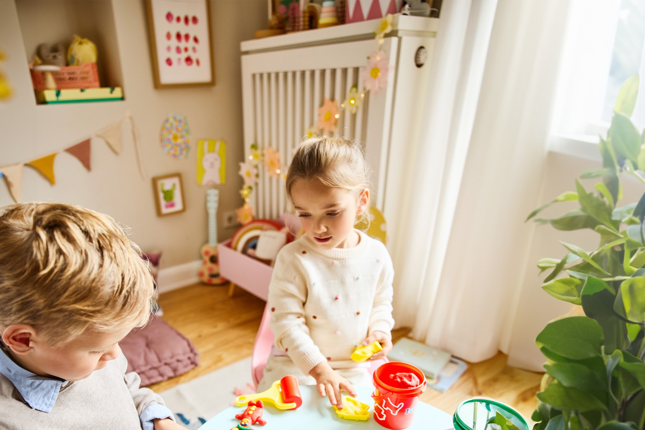 Bambini che giocano con pasta modellabile e accessori su un tavolo in una stanza luminosa.