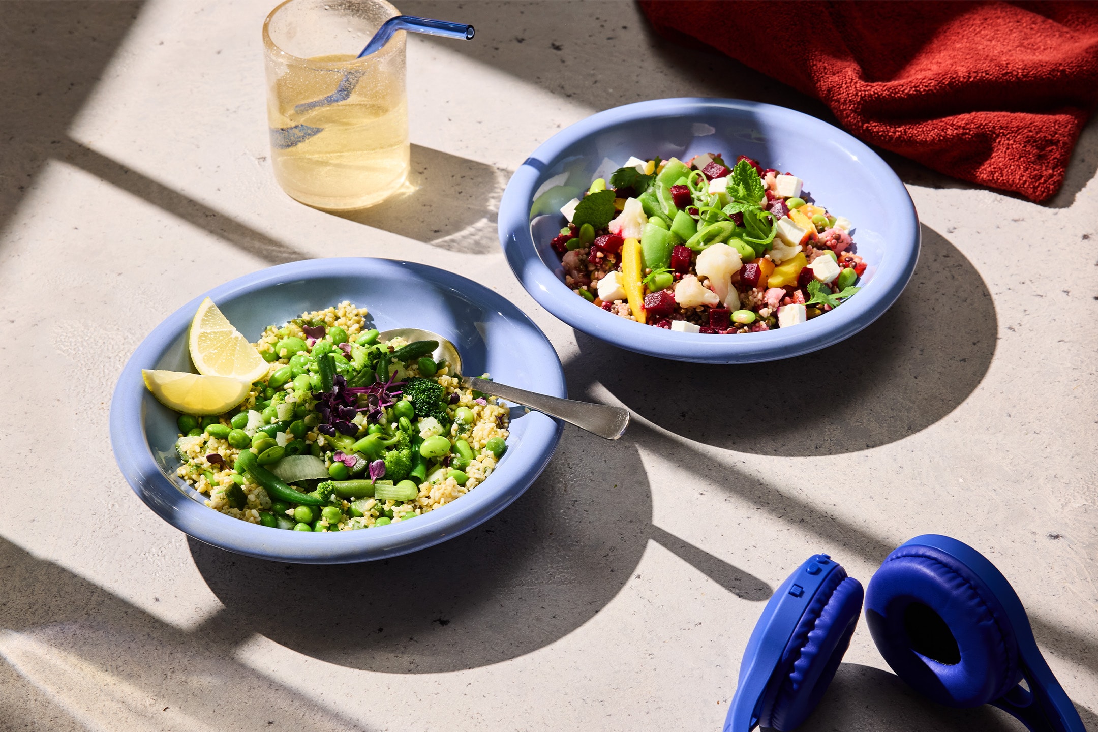 Zwei Schalen mit gesunden Salaten, ein Glas Wasser und blaue Kopfhörer auf einem Tisch.