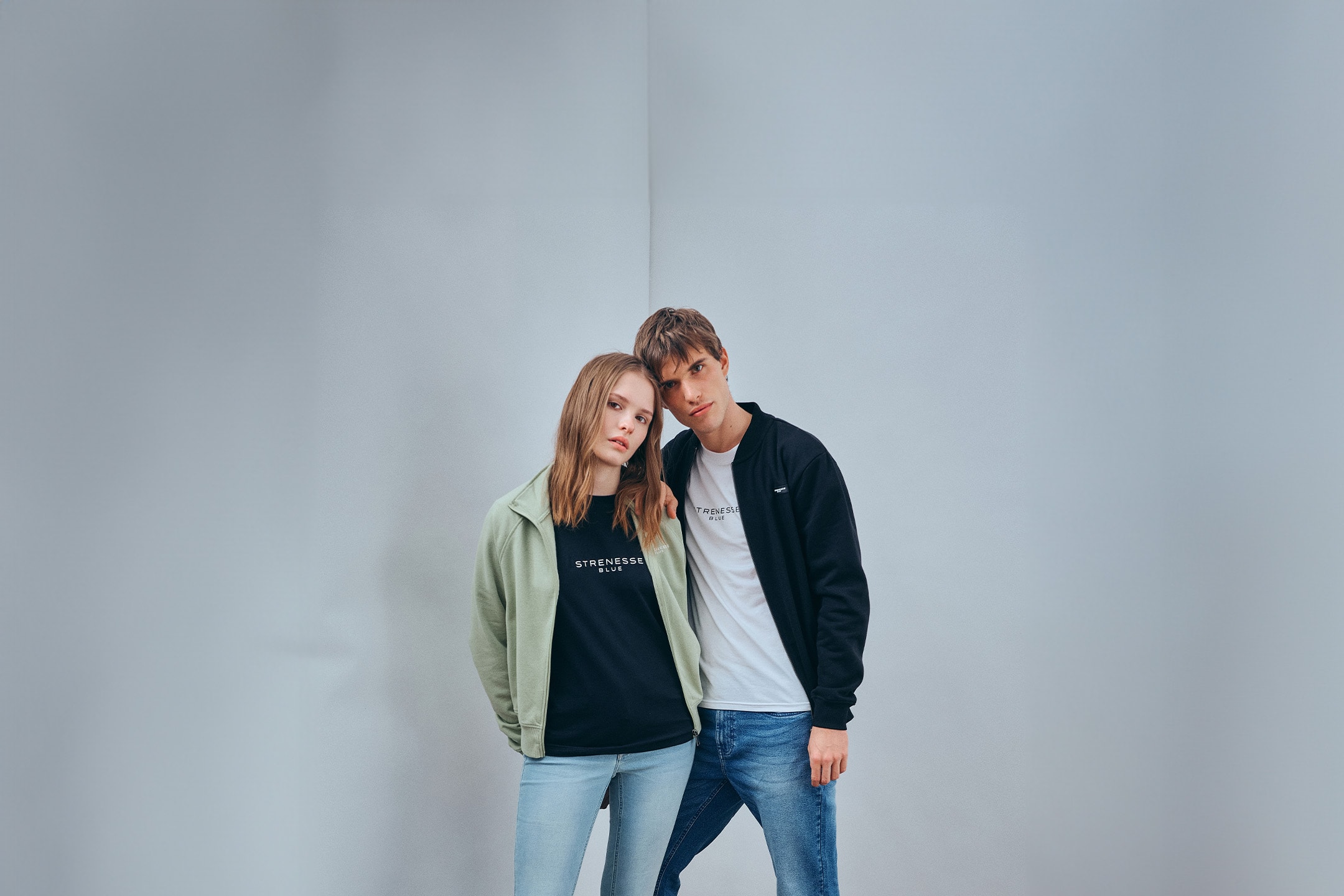Zwei Models tragen T-Shirts und Sportjacken mit Jeans in einem minimalistischen Studio.