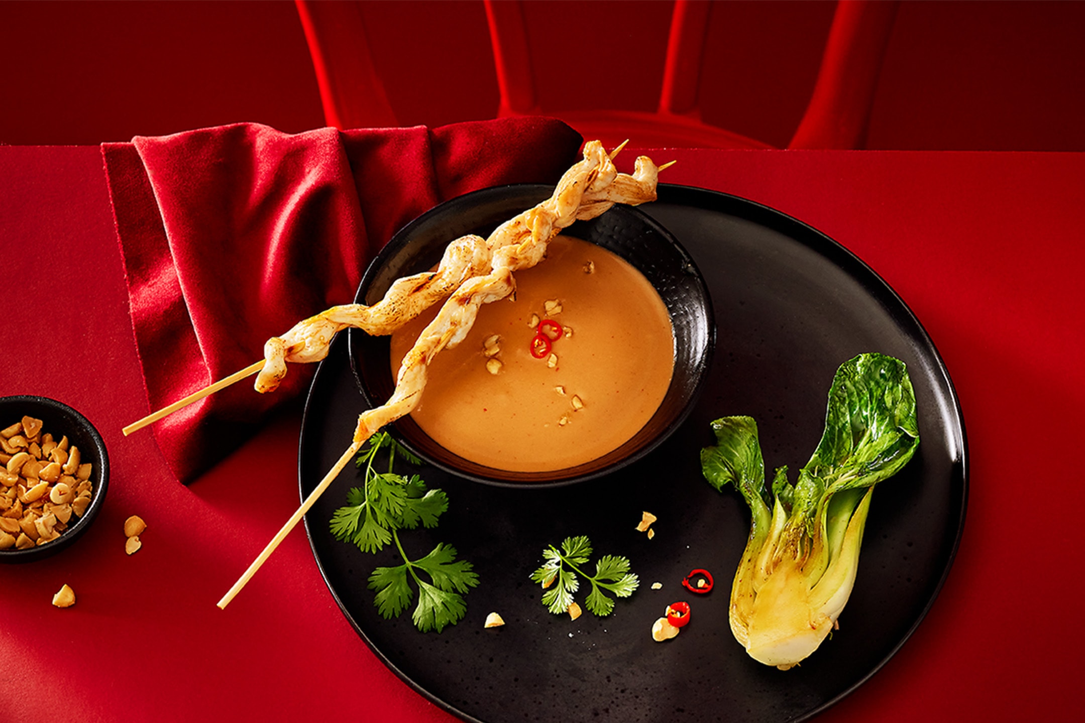 Satay-Hähnchenspieße mit Erdnusssauce, Pak Choi und Koriander auf rotem Hintergrund.
