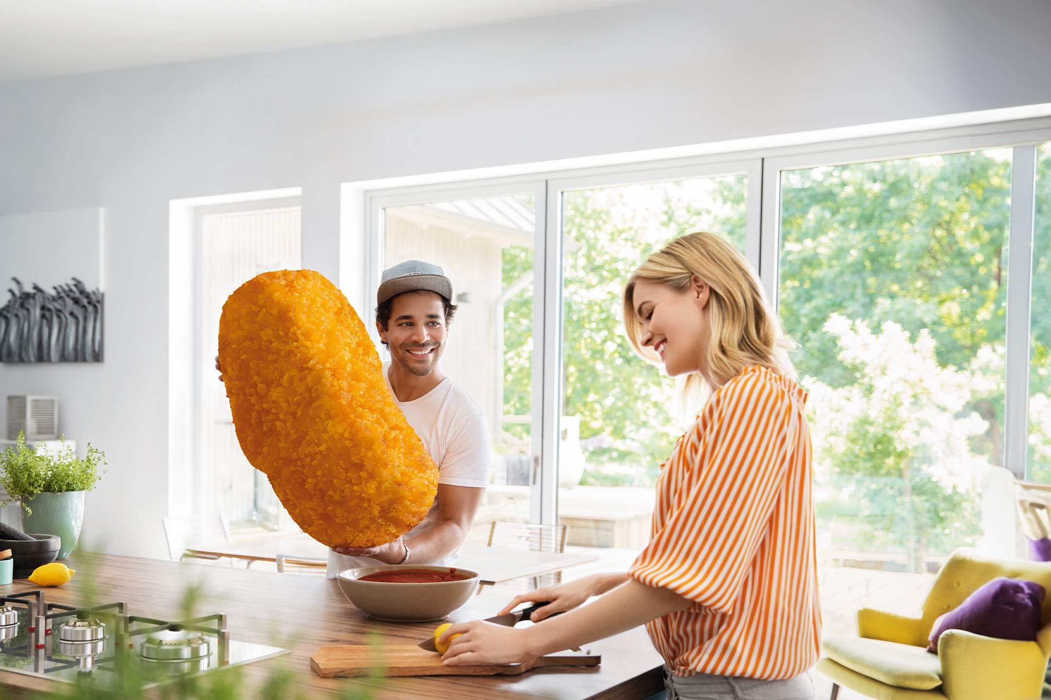 Un couple dans une cuisine, l'homme tient une énorme croquette, la femme coupe un citron.