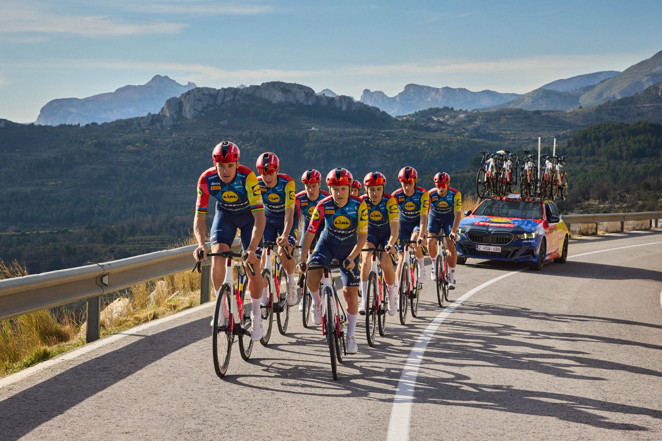 Radfahrerteam mit Begleitfahrzeug in bergigem Gelände, in farbenfroher Sportkleidung.