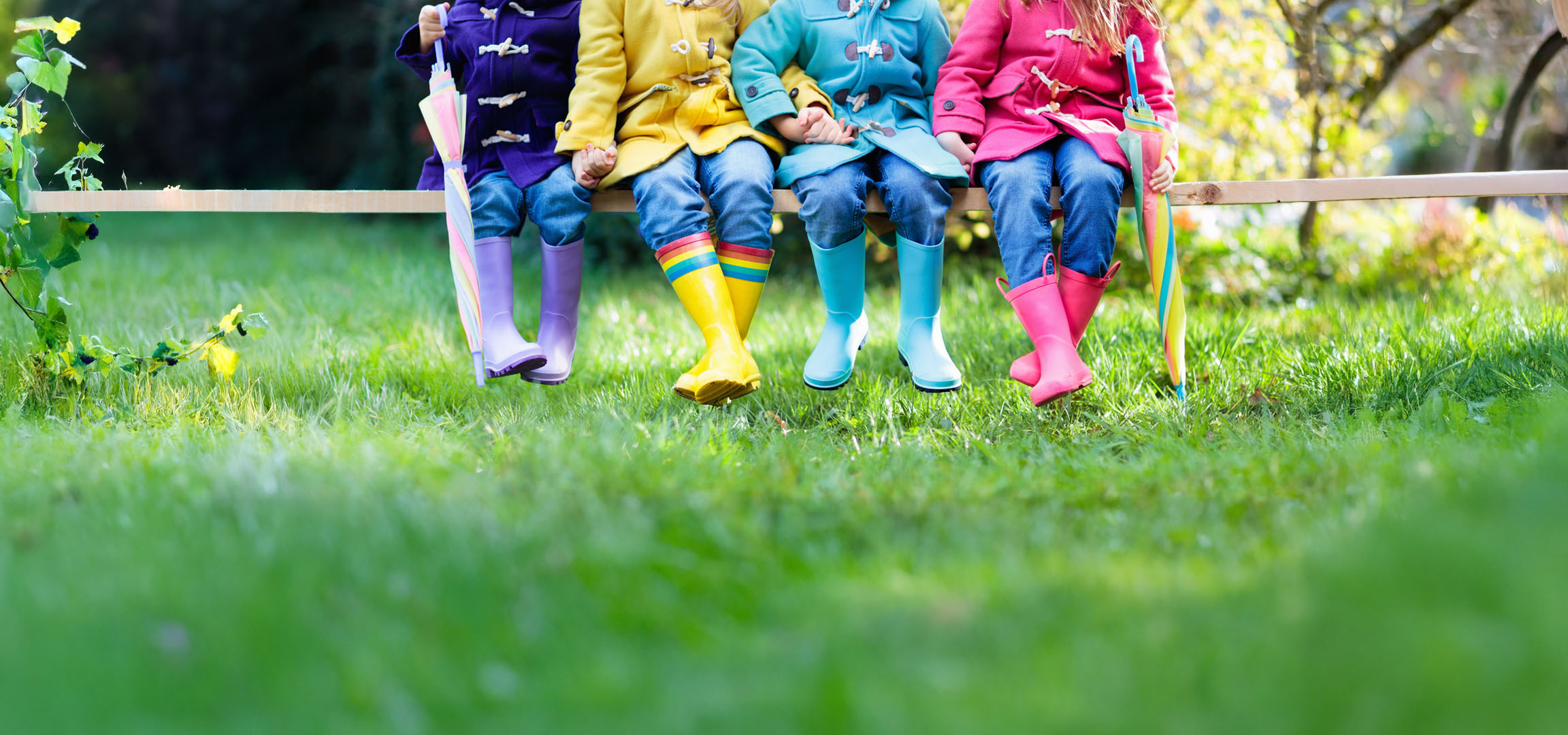 Enfants en imperméables et bottes colorés assis sur un banc dans l'herbe.
