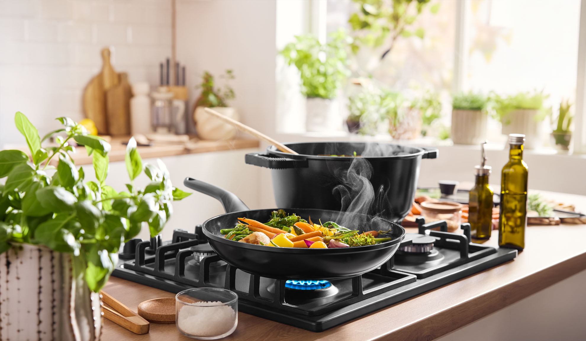 Dampfendes Gemüse kocht in Pfanne und Topf auf einem Gasherd in einer hellen Küche.