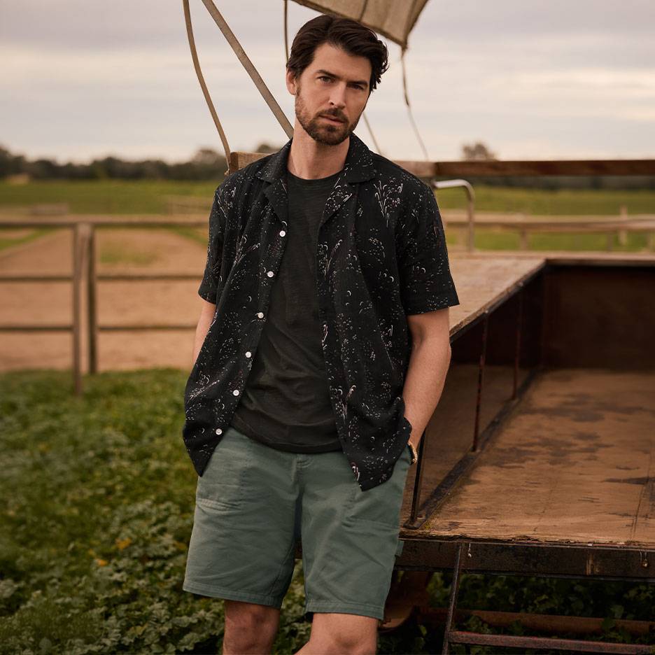 Homme en chemise noire à motifs, t-shirt et short vert, debout près d'une remorque.