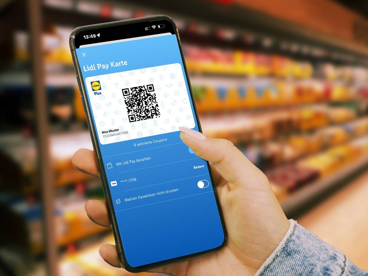 Eine Hand hält ein Smartphone mit der Bezahlkarte einer Supermarktkette, im Hintergrund sind Supermarktregale zu sehen.