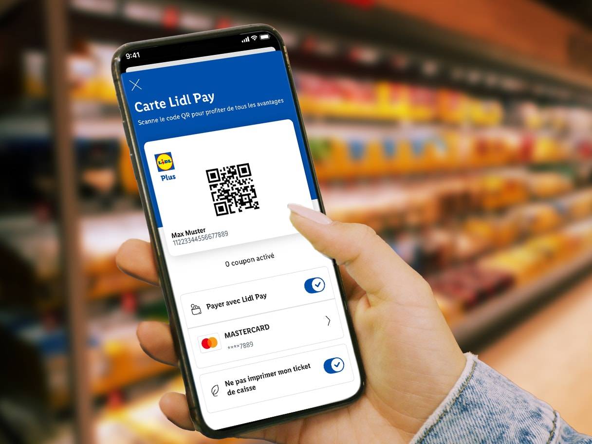 Une main tient un smartphone affichant l'application Lidl Pay avec un code QR et des options de paiement, devant un rayon de supermarché flou.