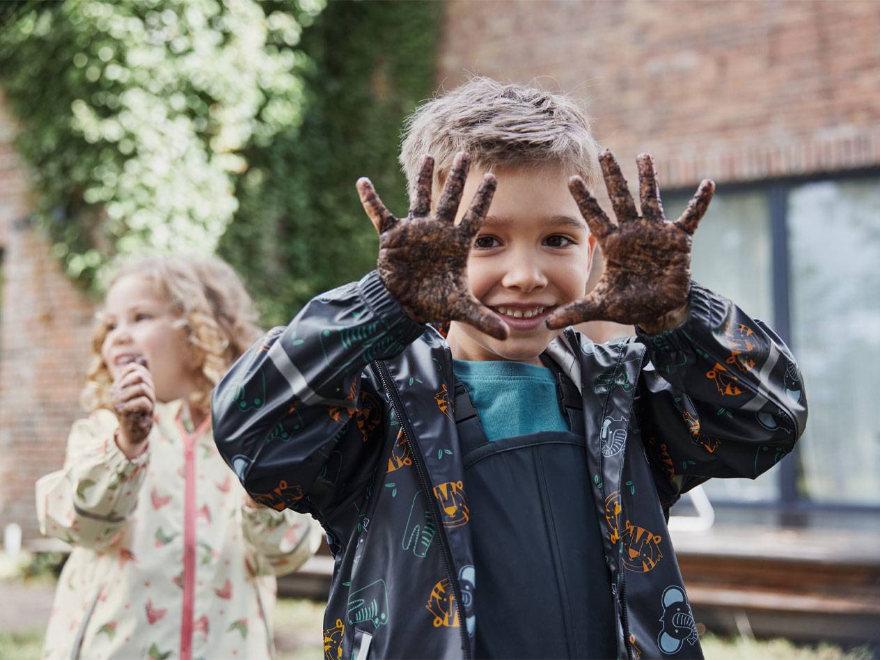 Deux enfants en vestes de pluie, les mains sales de boue, sourient à l'extérieur.