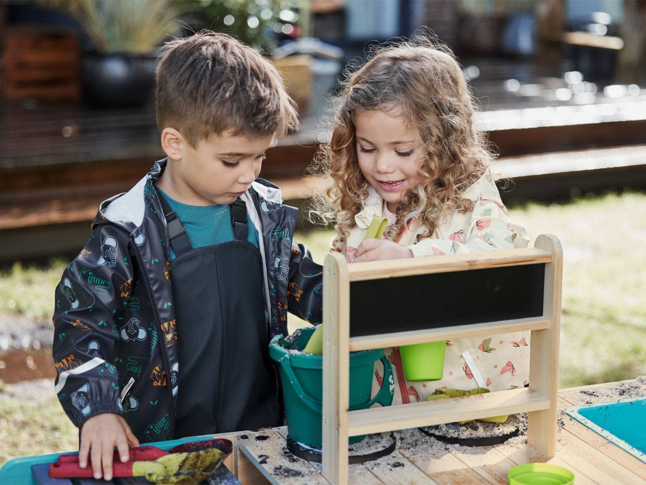Deux enfants en vestes de pluie et salopettes jouent avec du sable à une table de jeu.