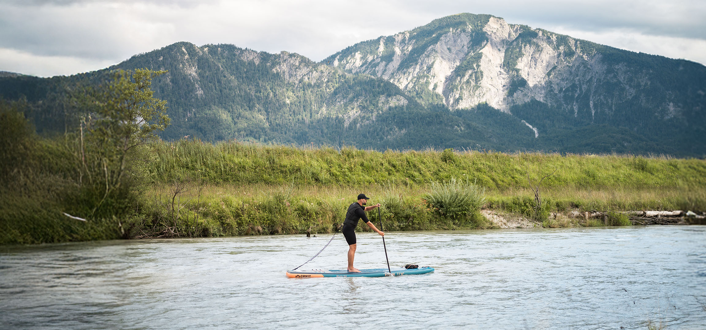 Uomo in paddleboard su un fiume con montagne e vegetazione lussureggiante sullo sfondo.