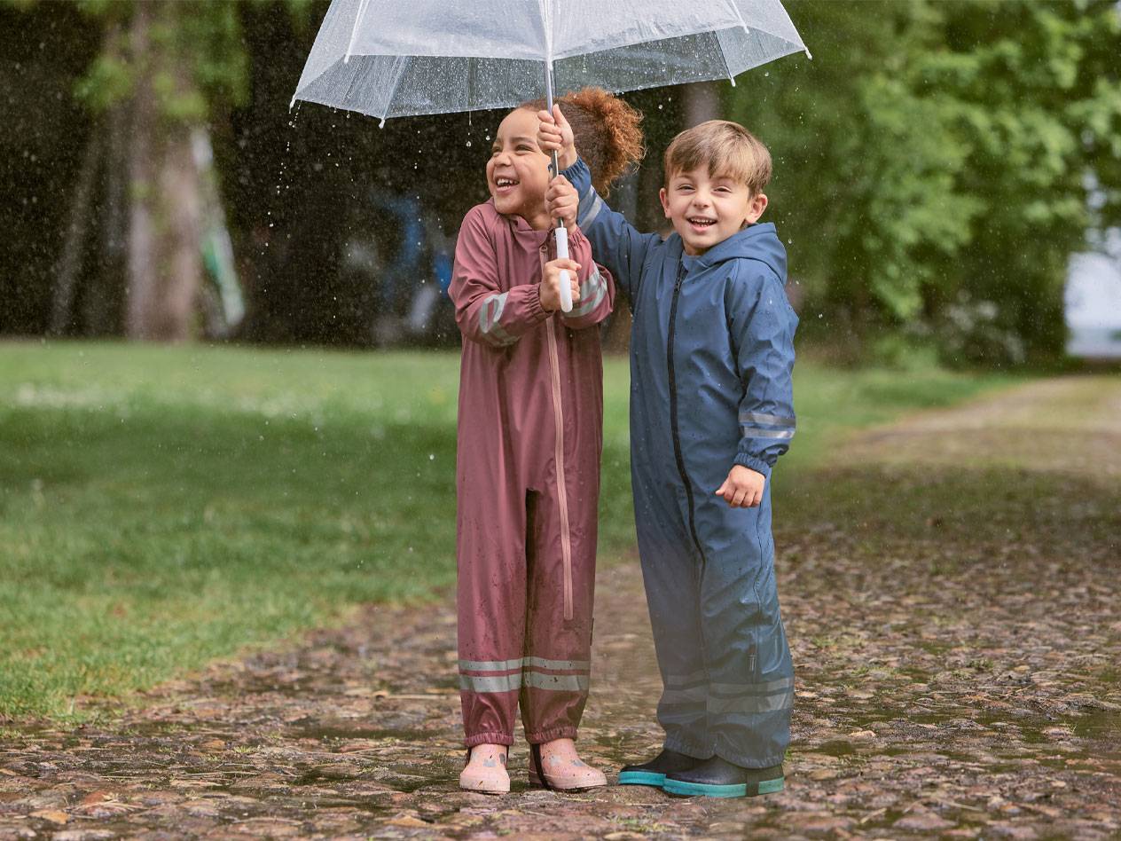 Deux enfants joyeux en combinaisons de pluie, sous un parapluie transparent.