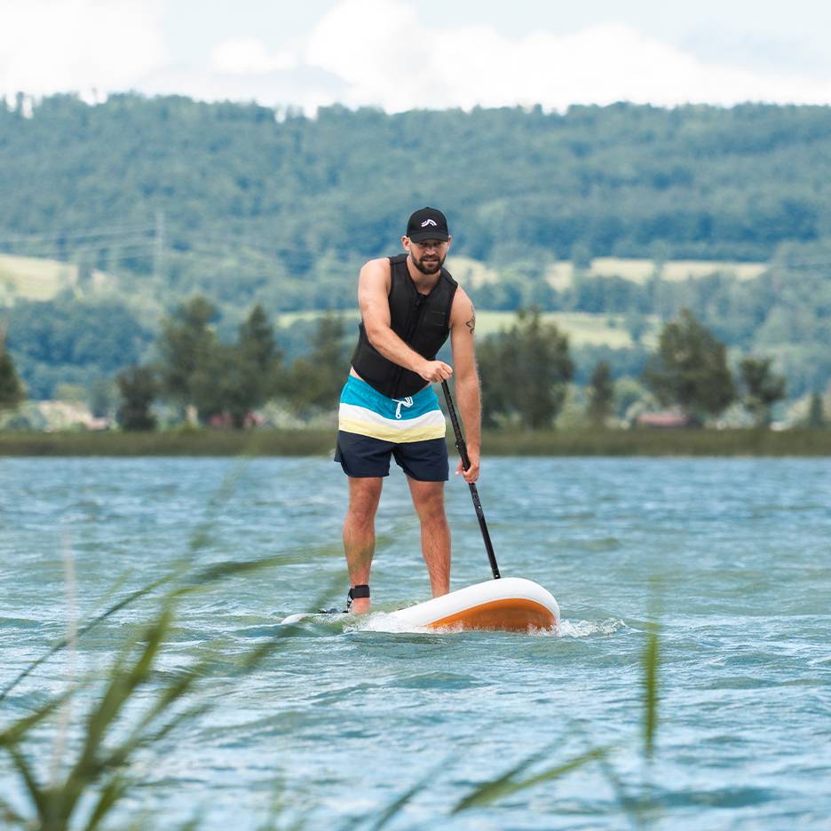 Uomo che fa stand-up paddle su un lago, con giubbotto di salvataggio e pantaloncini da bagno.