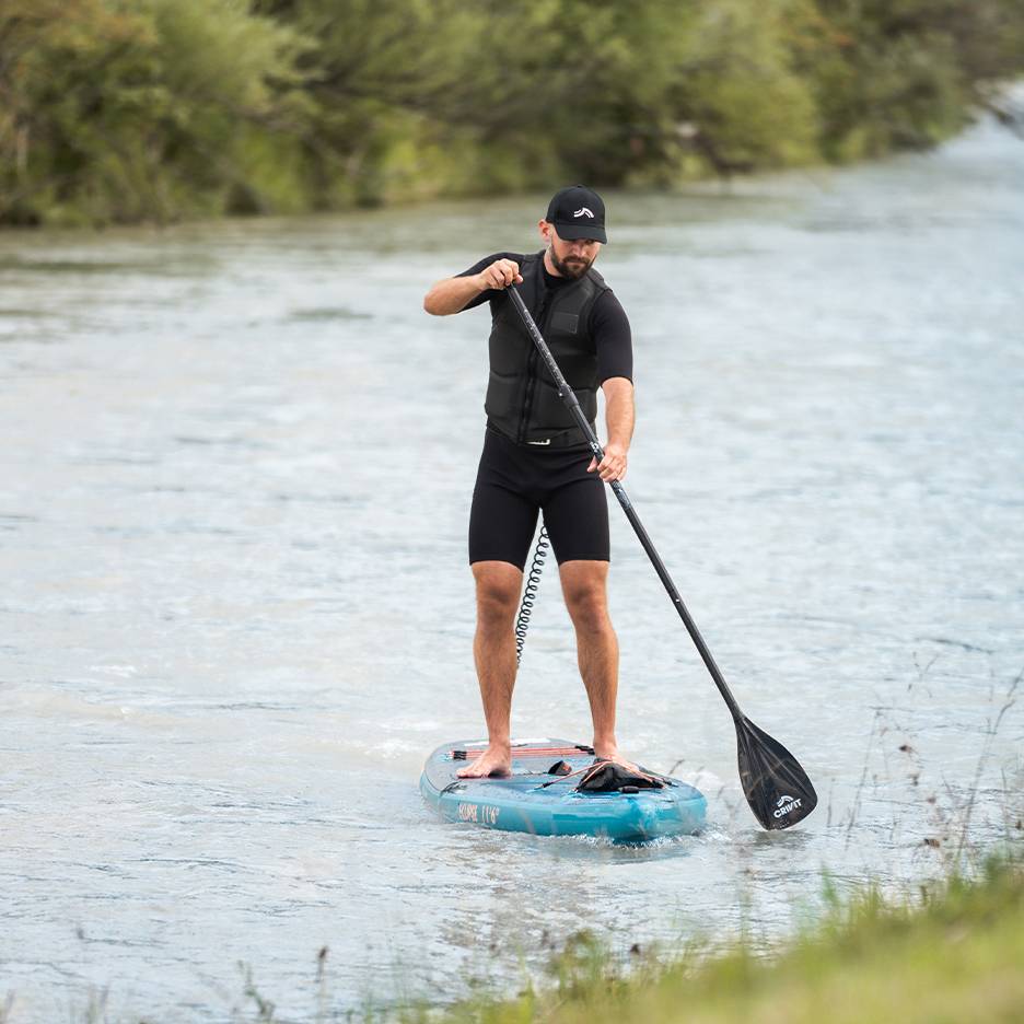 Uomo in paddleboard su un fiume, indossa muta nera, giubbotto di salvataggio e cappellino.