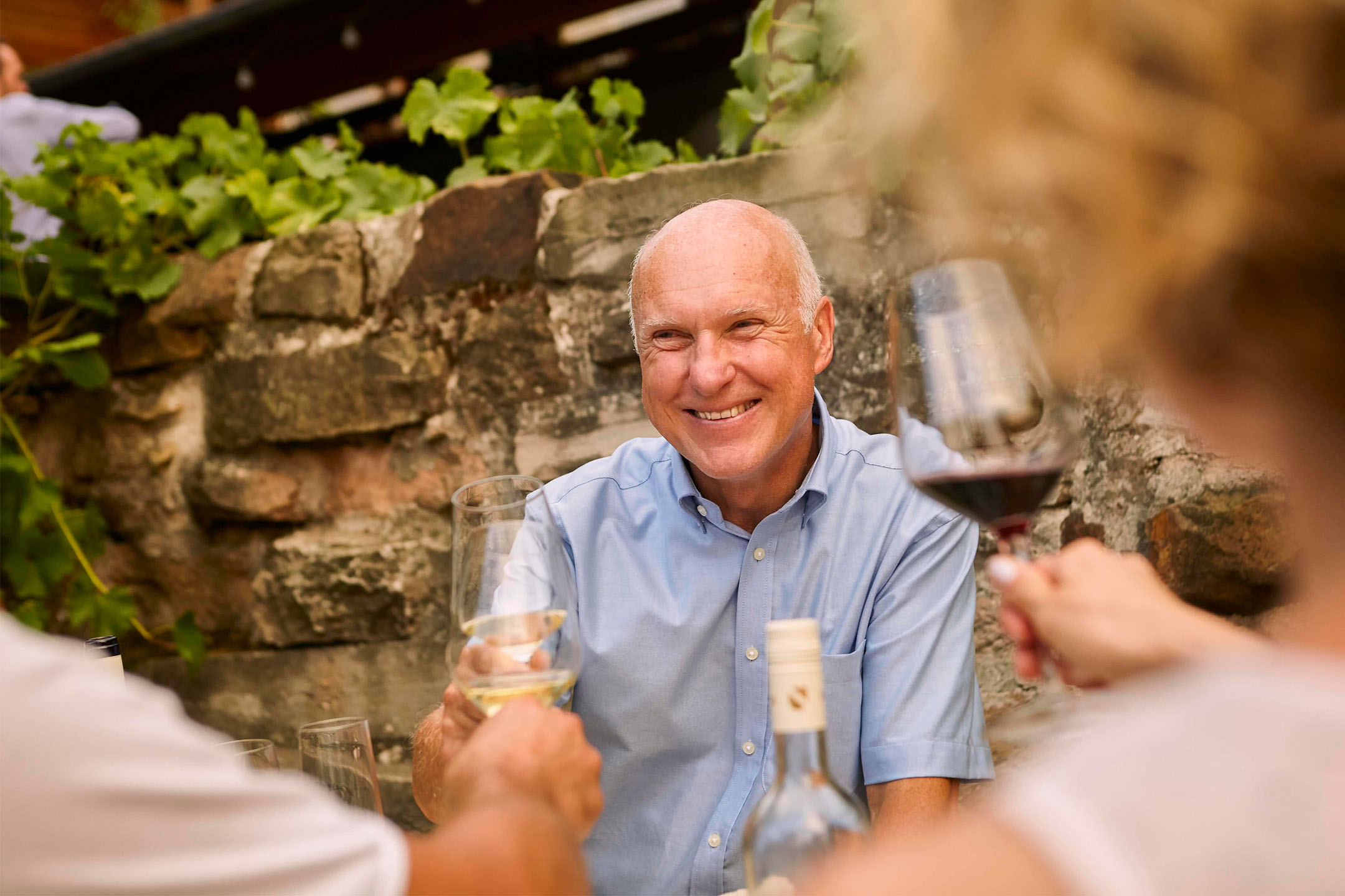 Homme souriant trinquant avec du vin blanc, entouré d'amis et d'un mur de pierre.