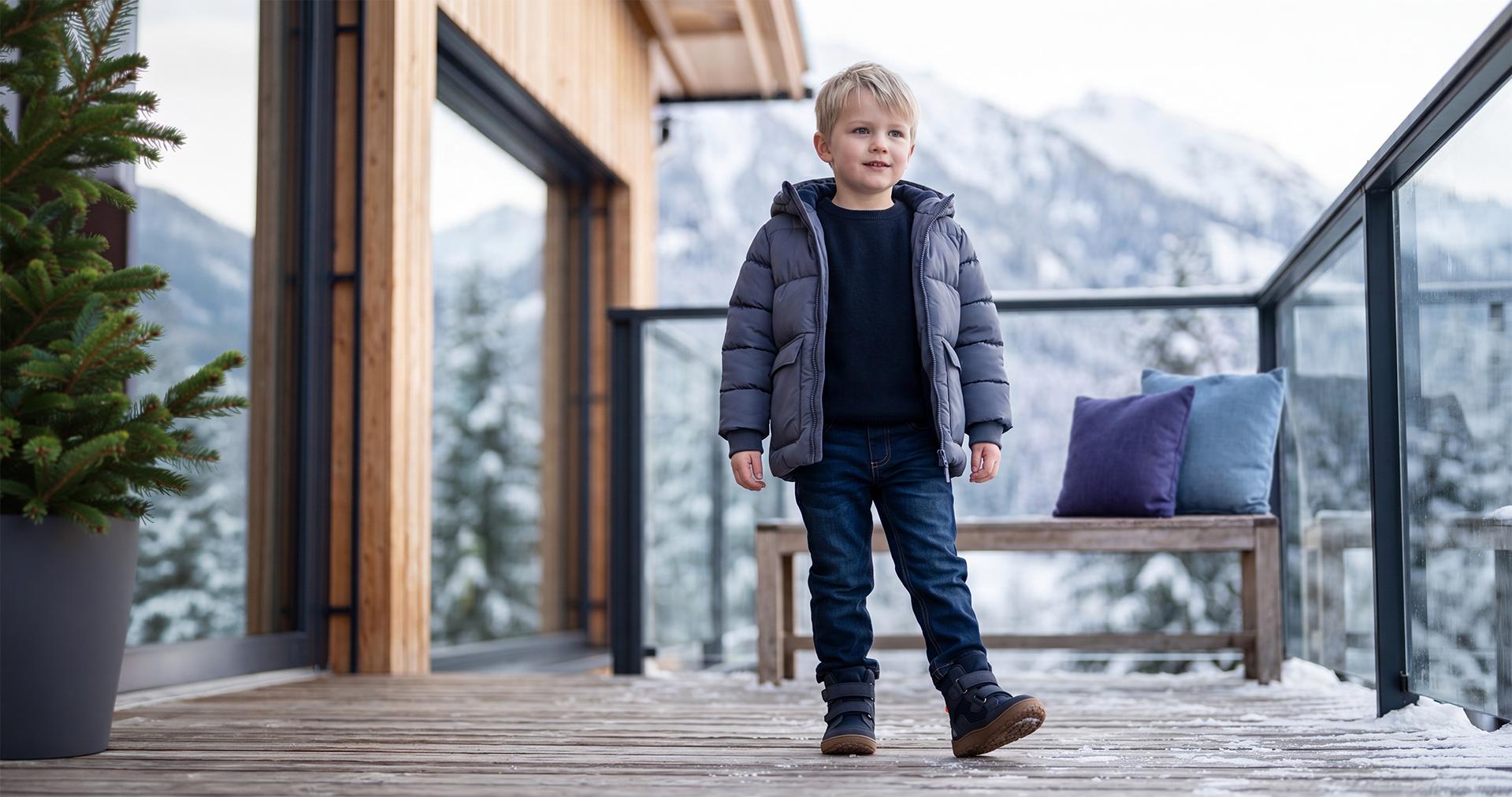 Kleiner Junge in warmer Winterjacke, Jeans und Stiefeln auf einer Bergterrasse