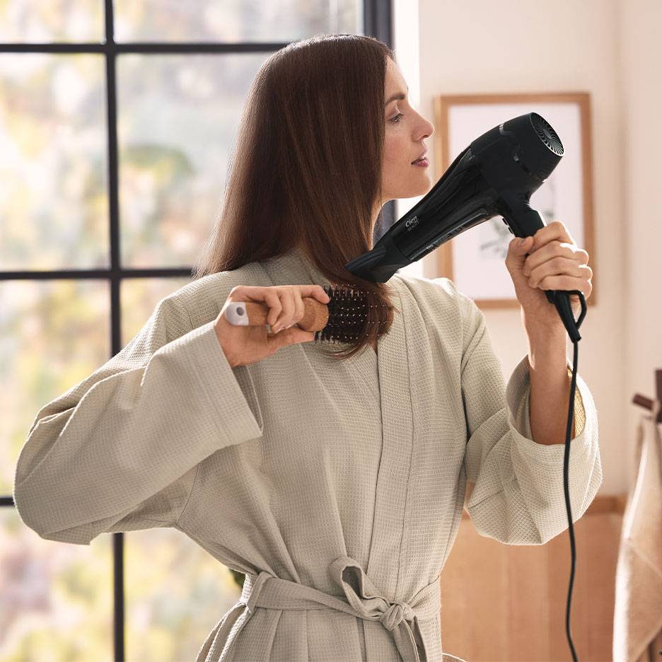 Femme se séchant les cheveux avec un sèche-cheveux et une brosse ronde, portant un peignoir.