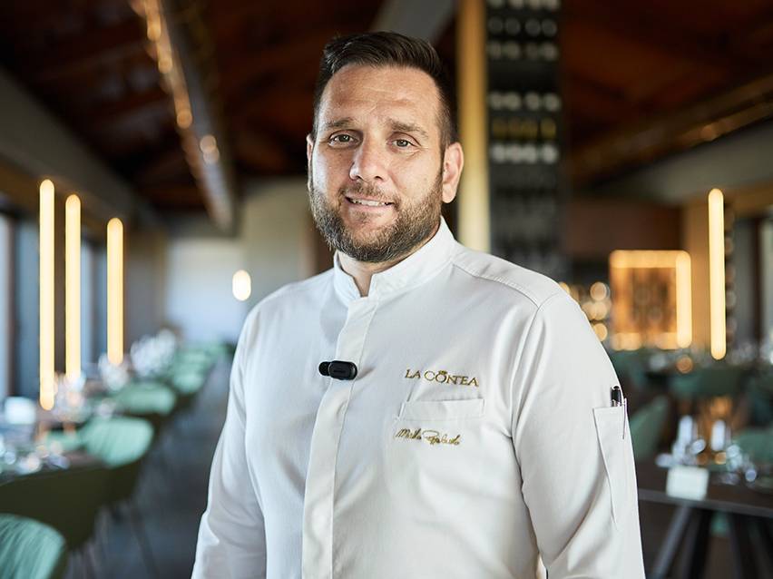 Chef souriant en uniforme blanc avec le logo 'LA CONTEA' dans un restaurant élégant.