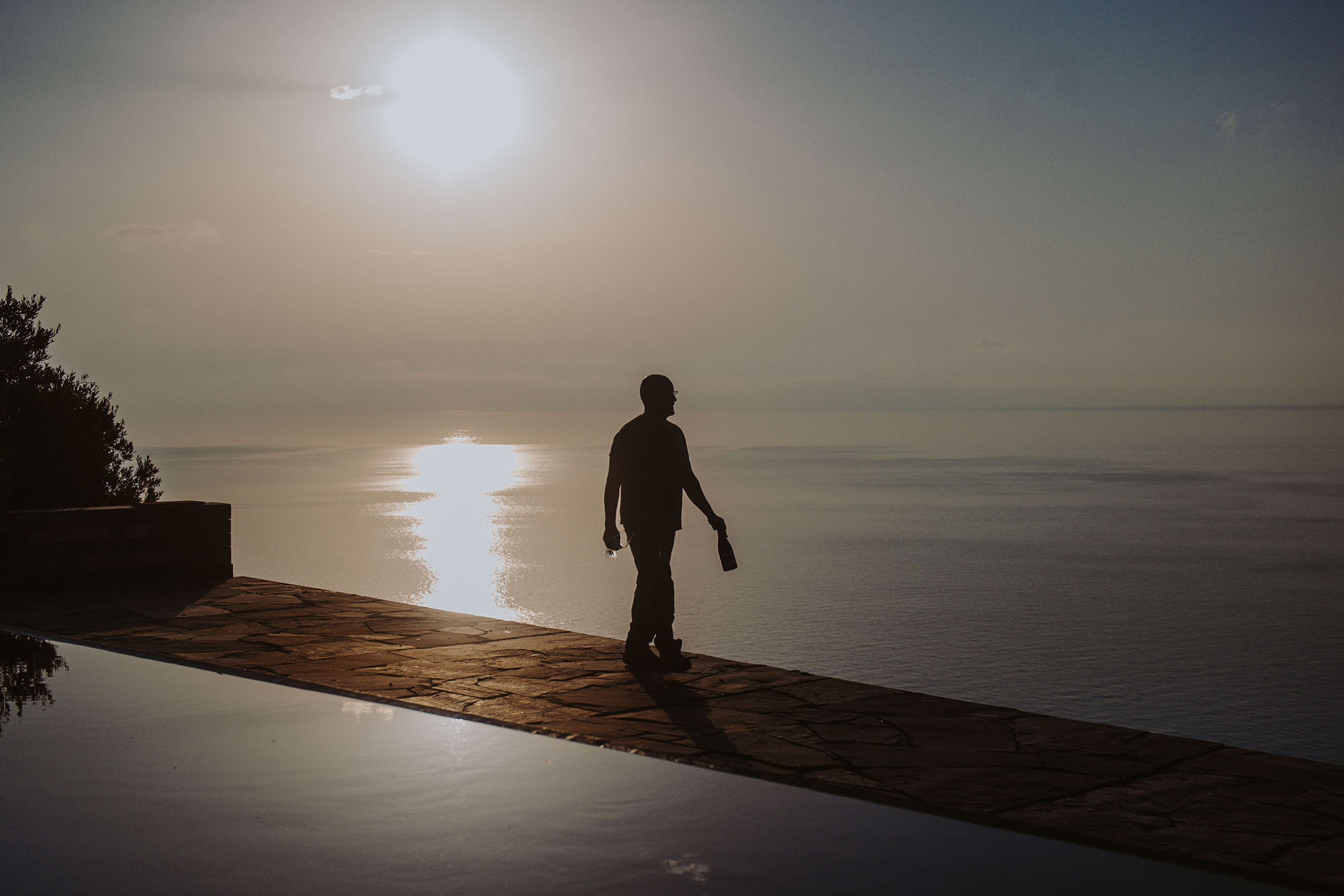 Silhouette d'une personne tenant une bouteille et un verre près d'une piscine à débordement au coucher du soleil