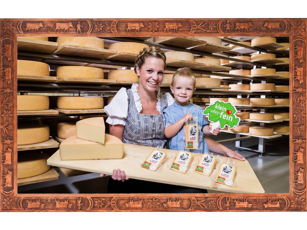 Donna e bambino presentano prodotti caseari davanti a scaffali di forme di formaggio, incorniciati da legno.