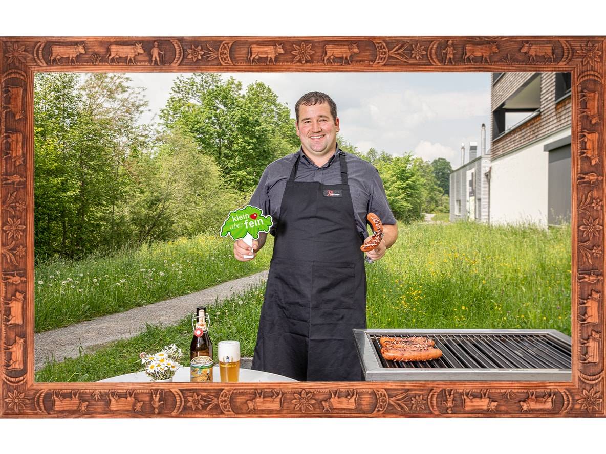 Uomo griglia salsicce, tiene un cartello 'klein aber fein' e una birra su un tavolo.