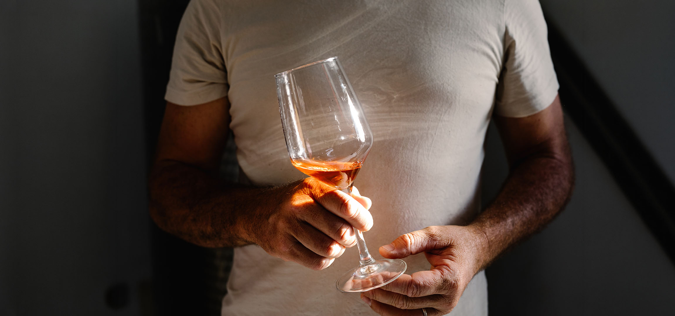 Mann hält ein Glas Roséwein, sonnenbeschienen, vor dunklem Hintergrund.