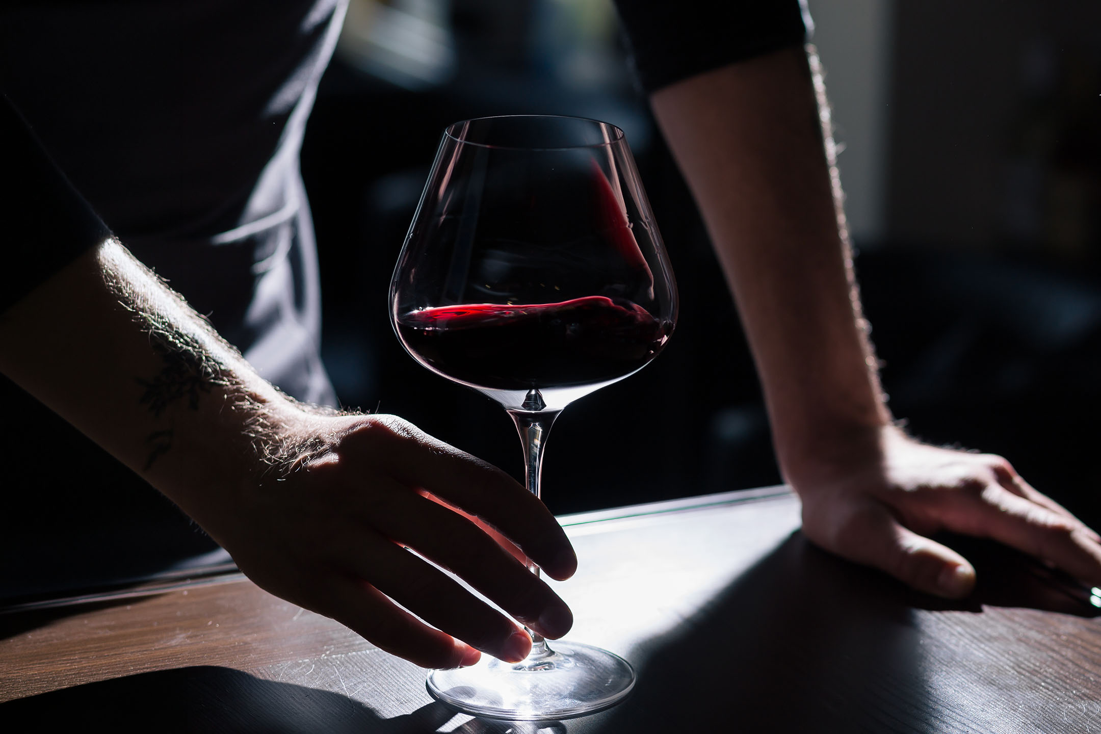 Tätowierte Hände eines Mannes halten ein Glas Rotwein auf einem Holztisch.