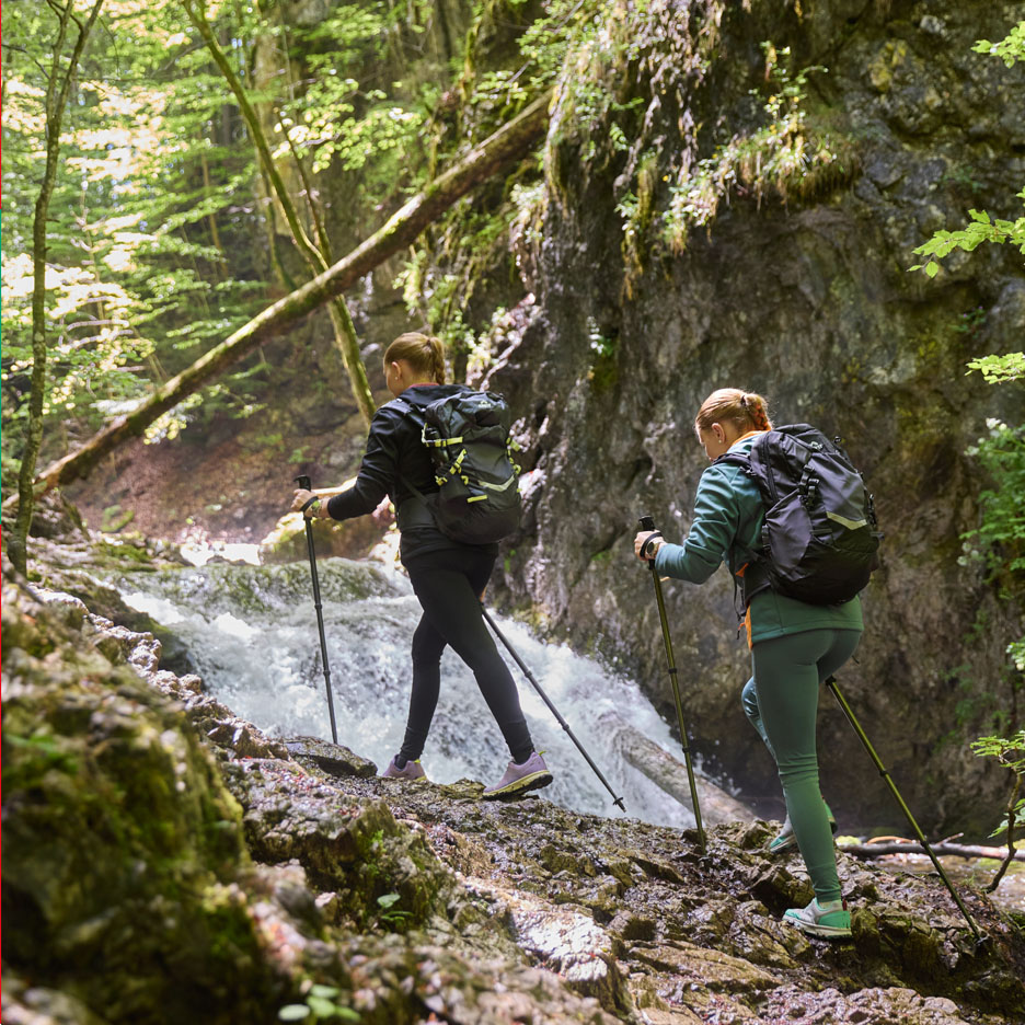 Due donne con zaini e bastoncini da trekking camminano lungo una cascata.