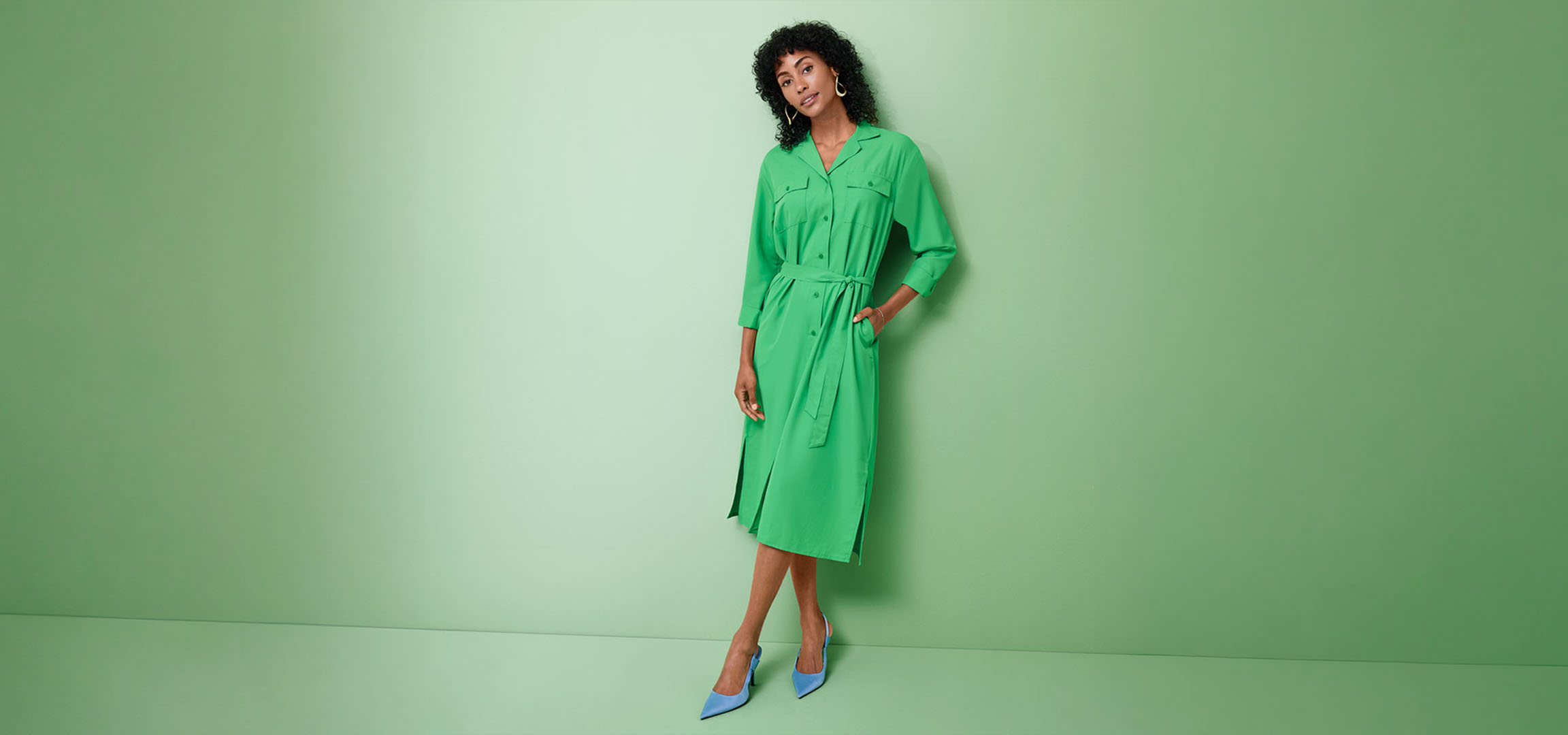 Femme en robe chemise verte et talons bleus, posant sur un fond vert.