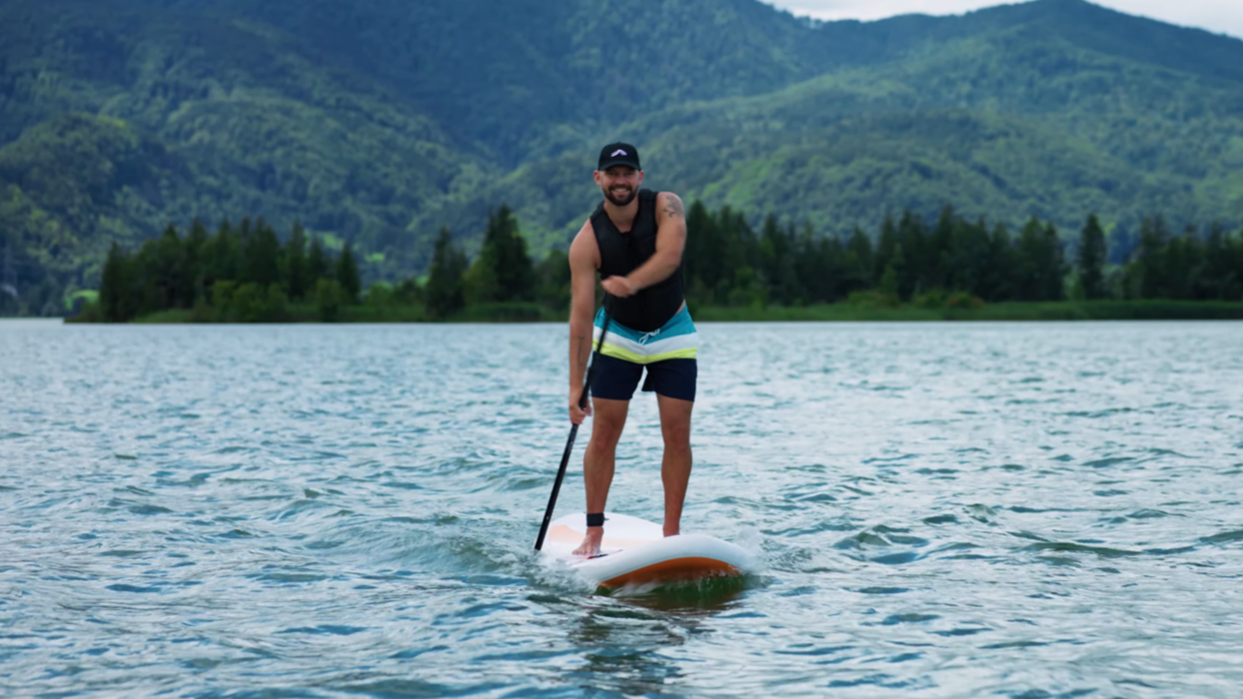 Mann auf Stand-Up-Paddle-Board auf einem See mit Bergen im Hintergrund.
