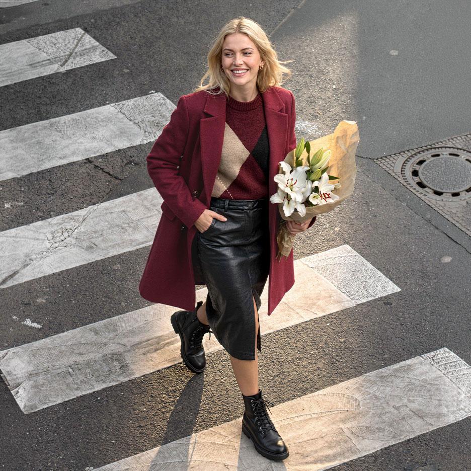Femme souriante en manteau bordeaux, pull argyle et jupe en cuir, traversant la rue avec un bouquet de lys.