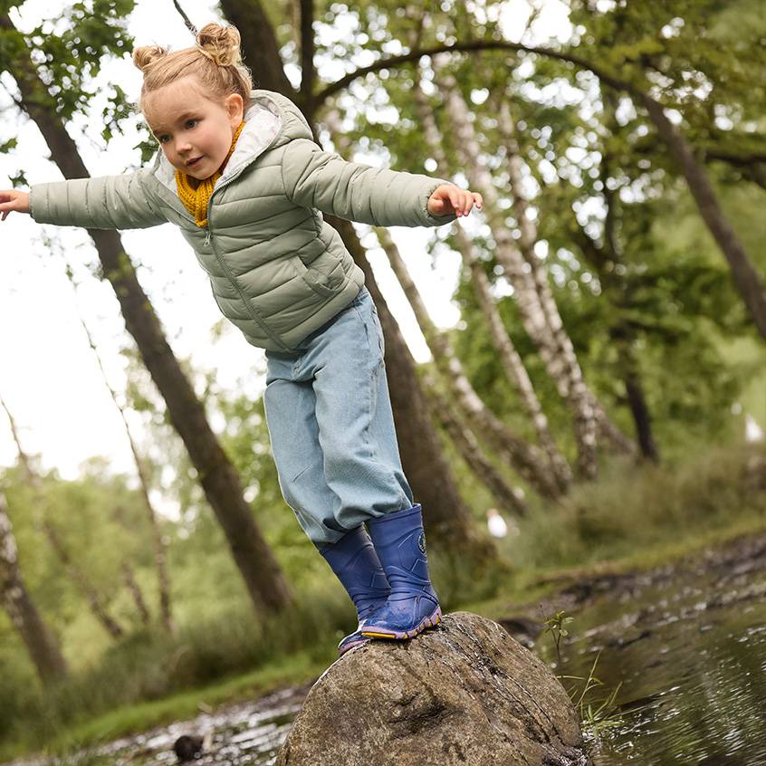 Petite fille en doudoune, jean et bottes en caoutchouc en équilibre sur un rocher dans les bois.